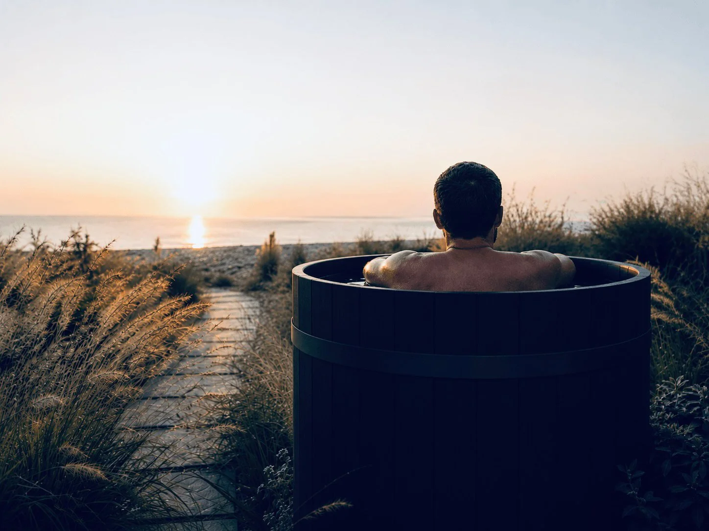 A person sitting in a hot tub facing a sunset over the ocean, with a pathway surrounded by plants leading towards the beach.