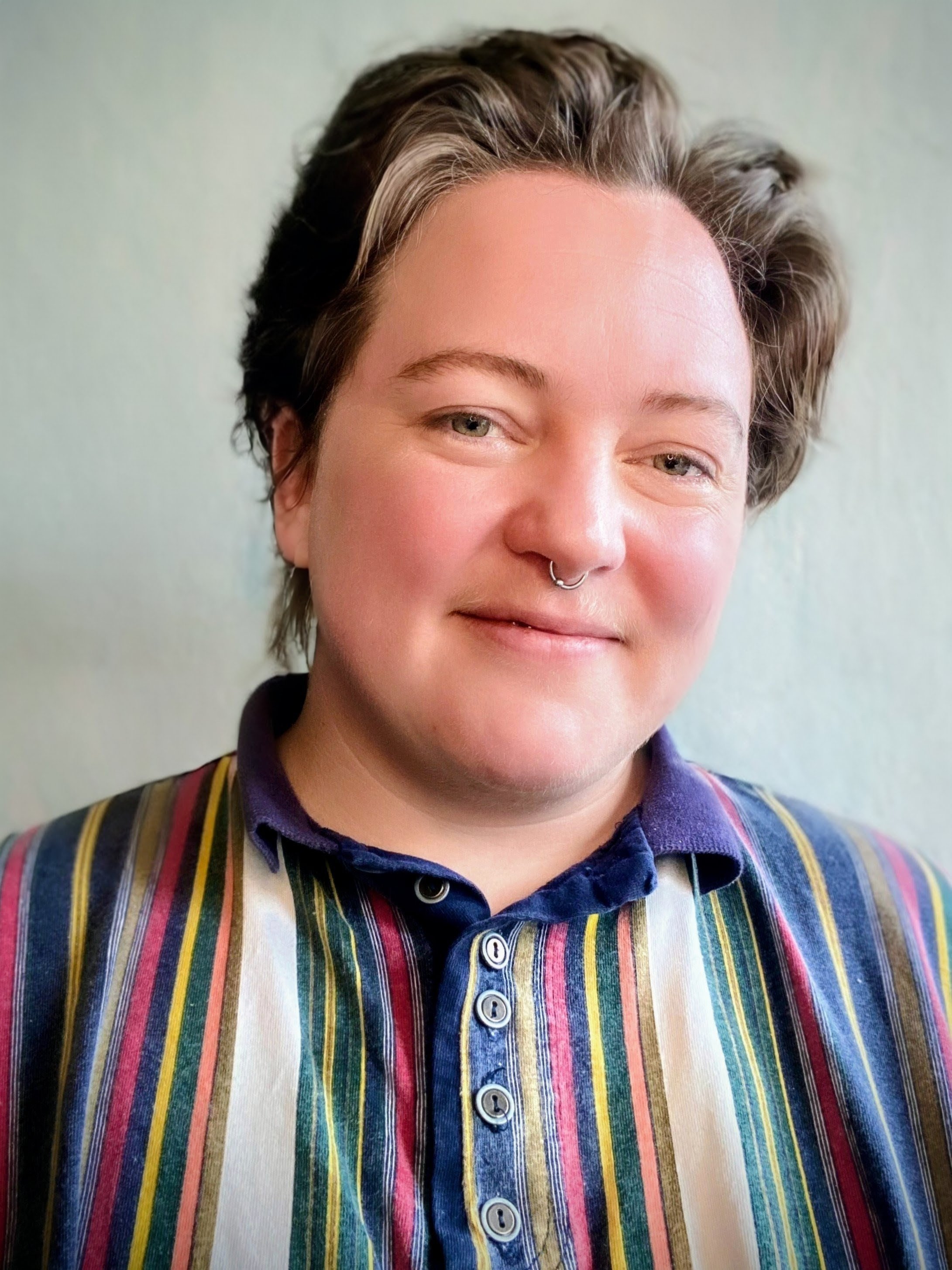 Close-up of a person with short, wavy hair, wearing a colorful striped shirt, smiling, with a septum piercing and light-colored eyes.