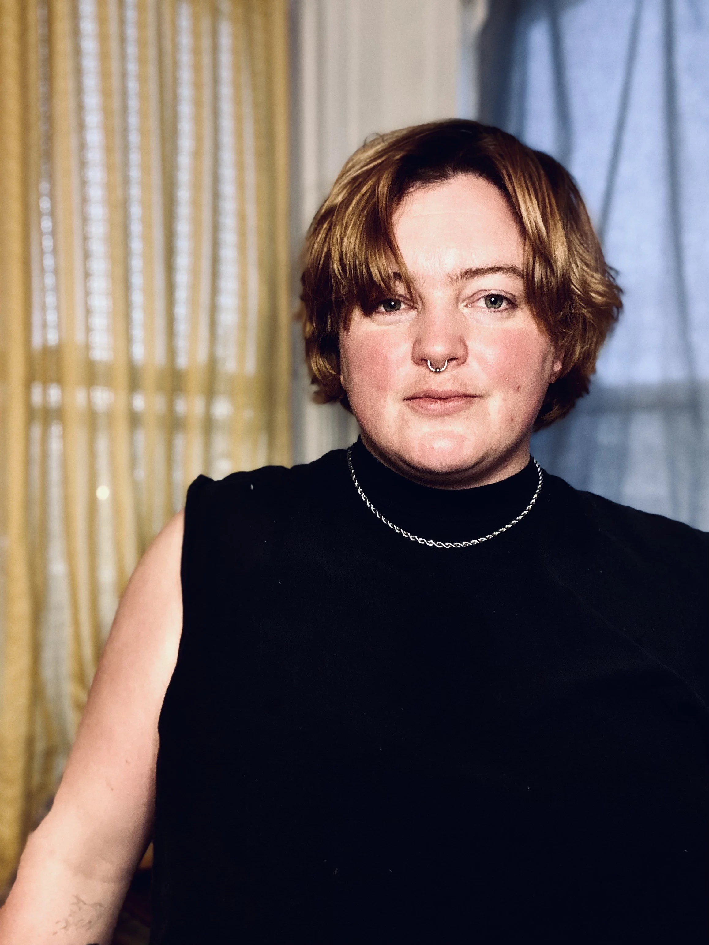 Person with short brown hair and septum piercing wearing a black sleeveless top and silver chain necklace, standing indoors with beige curtain and blue curtain in the background.
