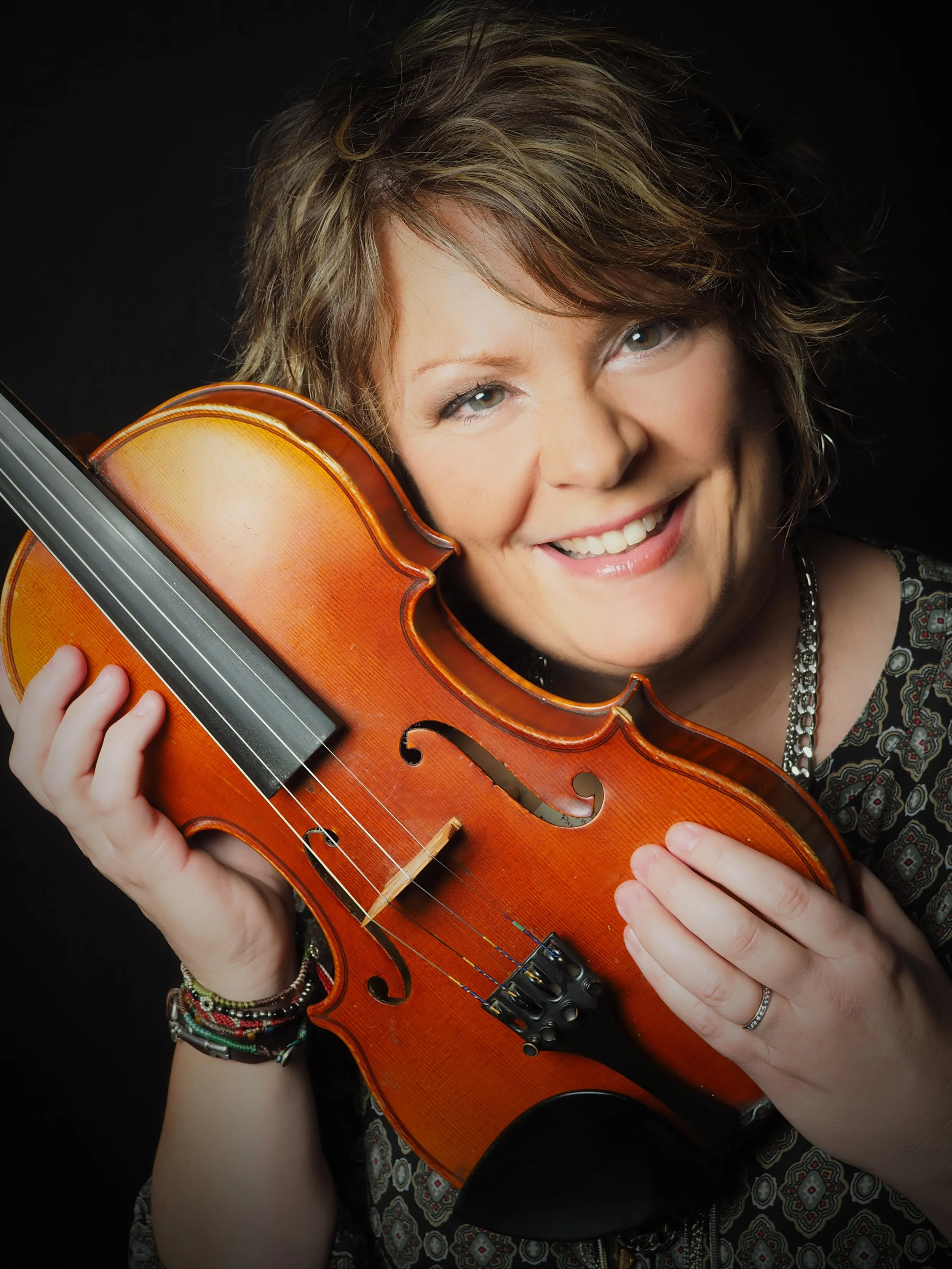 A woman with short, wavy hair smiling and holding a violin close to her face against a black background.