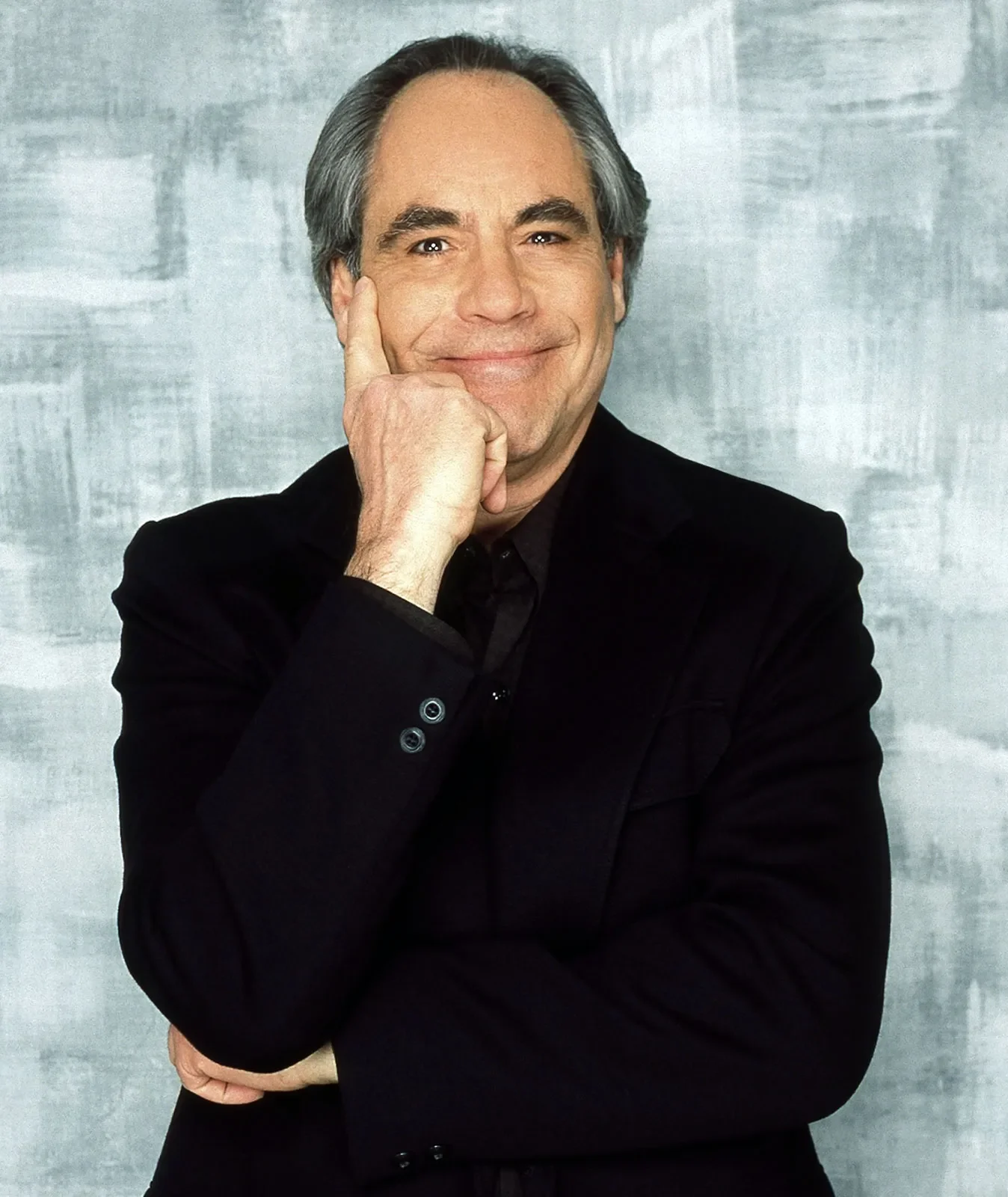 A middle-aged man with gray hair, dressed in a black suit, is smiling while resting his chin on his right hand, with his left arm crossed over his body, against a textured gray background.