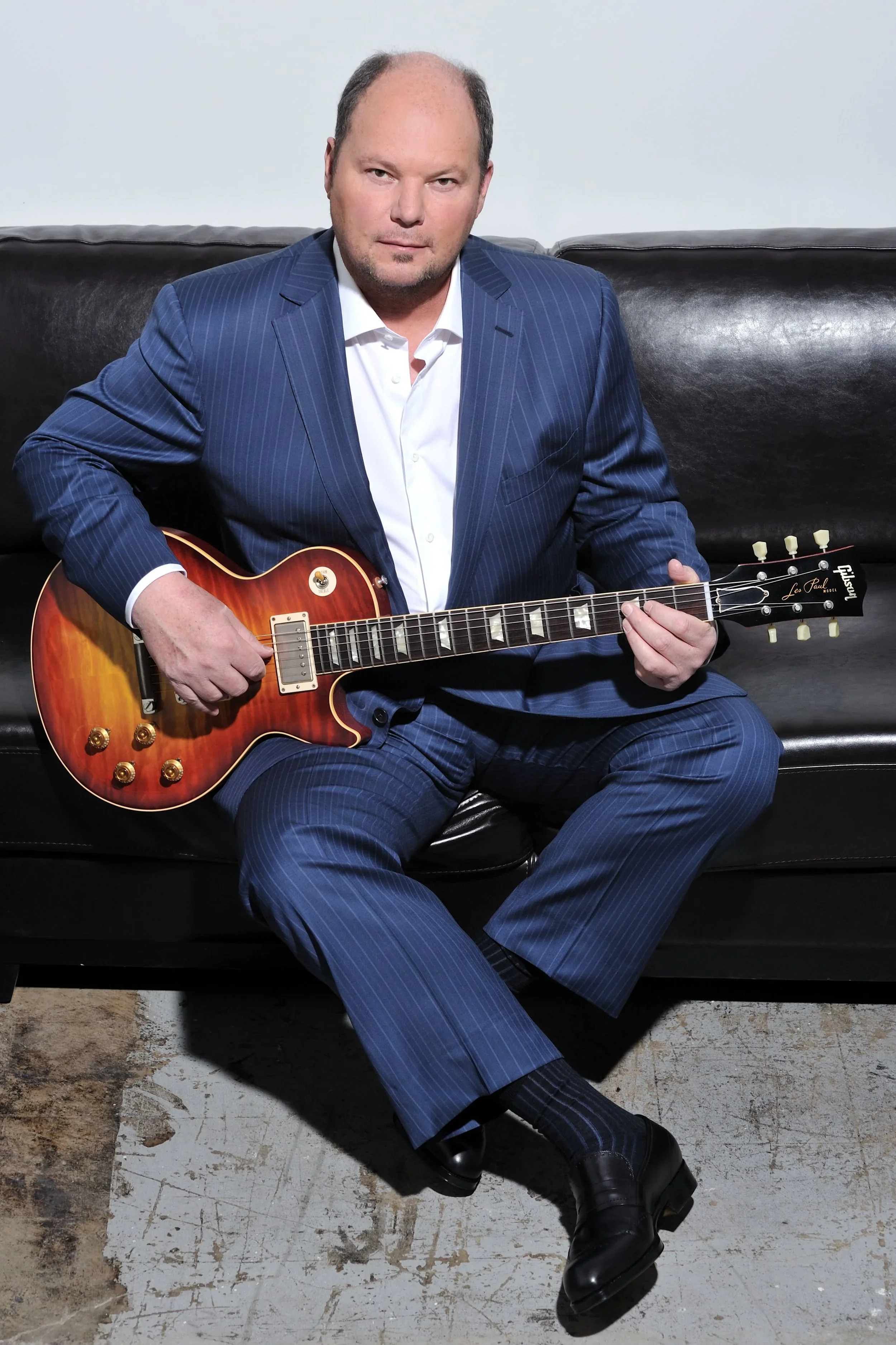 A man in a blue pinstripe suit sitting on a black leather couch, playing an orange Gibson Les Paul electric guitar, with his legs crossed and wearing black shoes, in a room with a worn concrete floor.