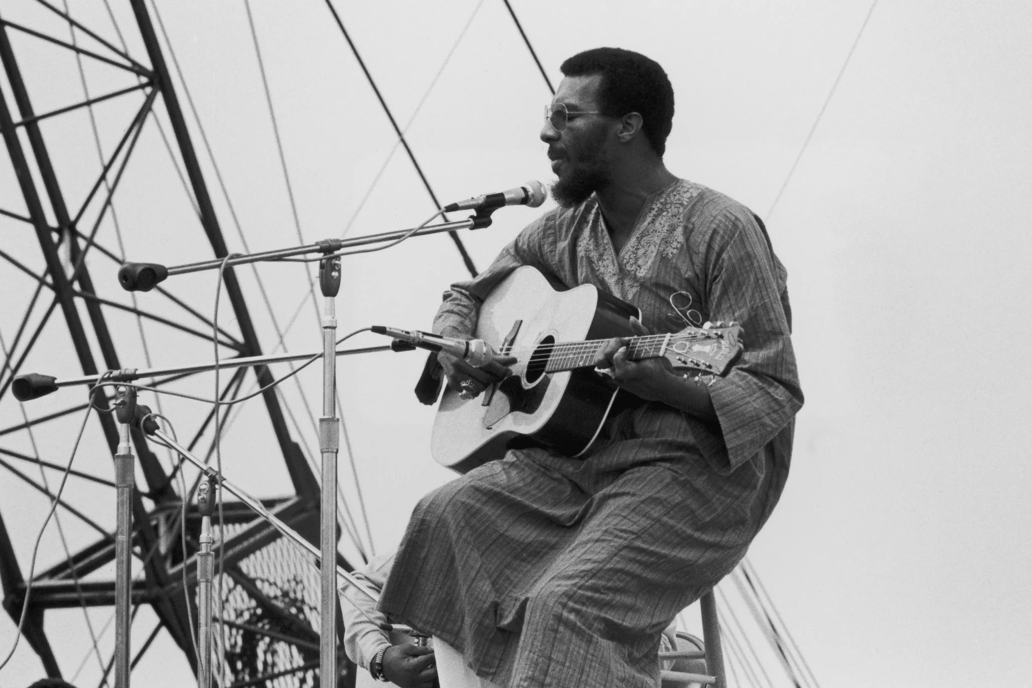 A man with dark skin, glasses, and a beard playing an acoustic guitar and singing into a microphone at an outdoor stage surrounded by metal structures.