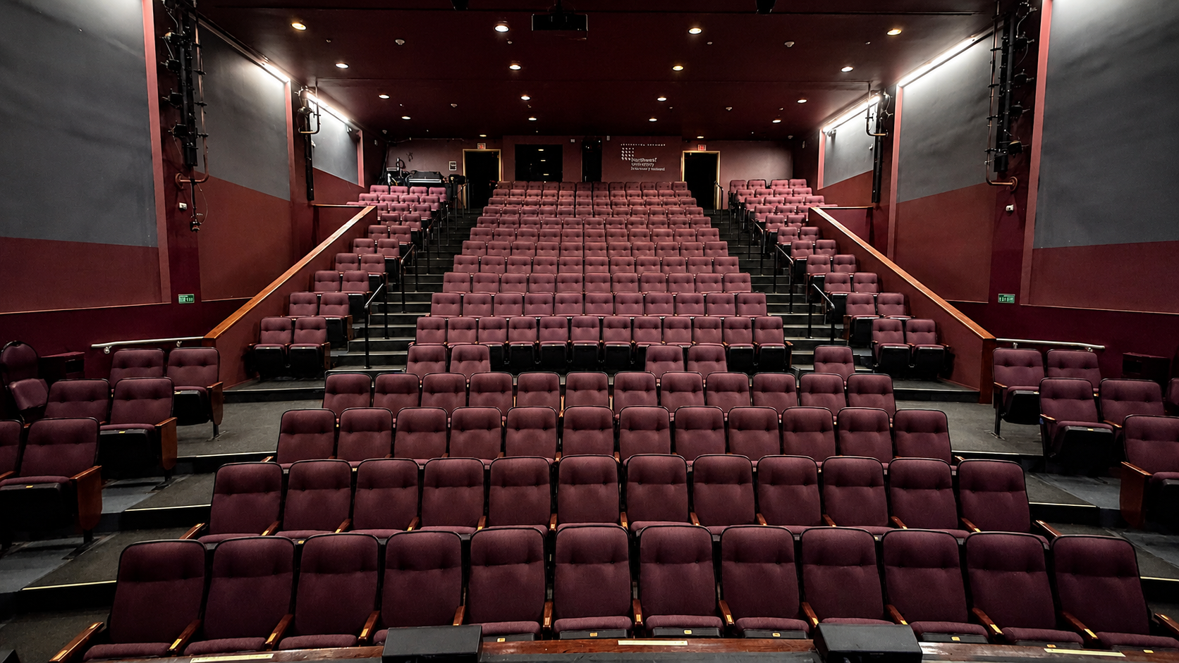 Empty theater with multiple rows of maroon cushioned seats, dark walls, and a stage at the front.