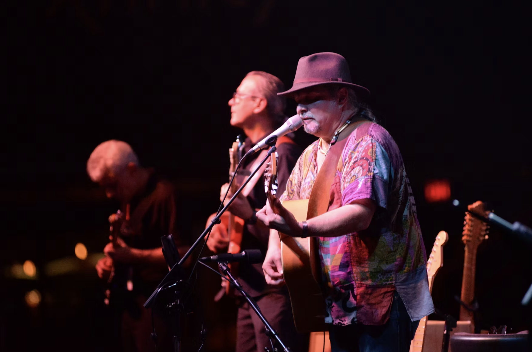 Three musicians performing on stage, with the one in the foreground wearing a patterned shirt, a hat, and playing an acoustic guitar. The other two in the background are also playing guitars.