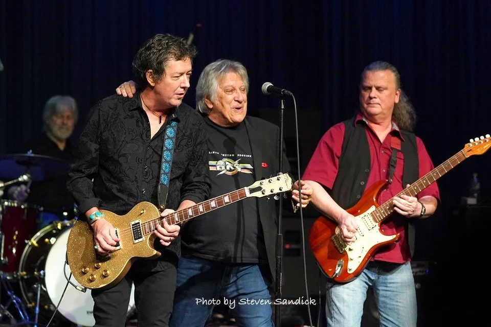 Four musicians performing on stage: one playing guitar, one singing into a microphone, one playing electric guitar, and a drummer in the background. The band appears to be performing live in a darkened venue.