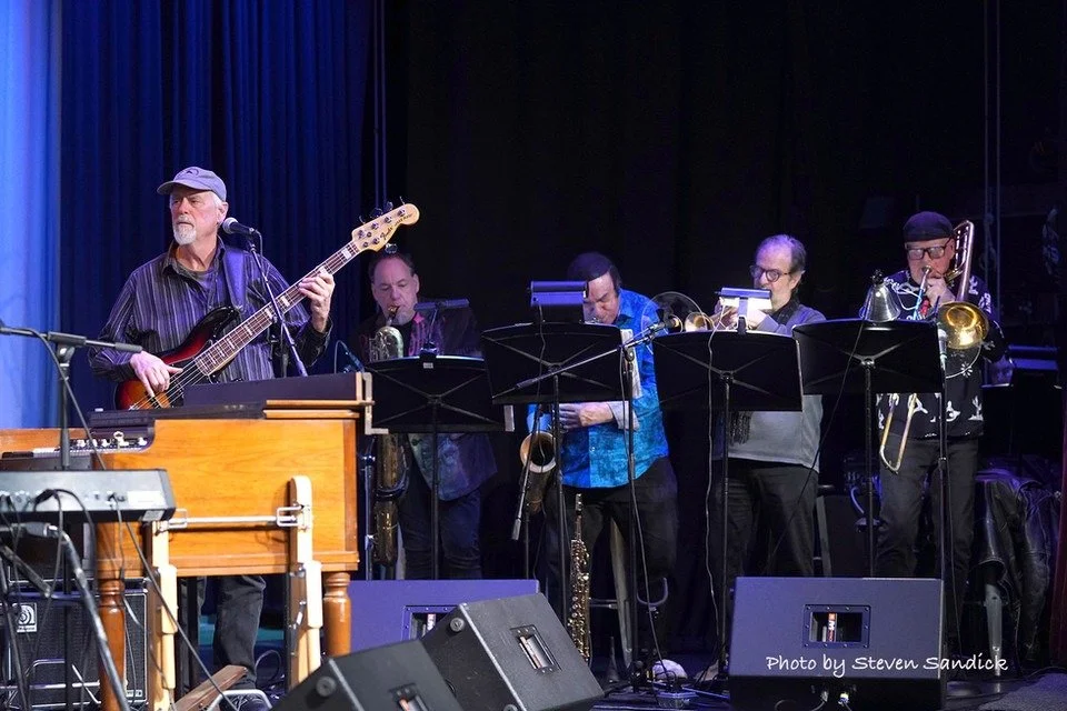 A band performing on stage with a guitarist and four saxophonists, with music stands and microphones, in a dark venue.