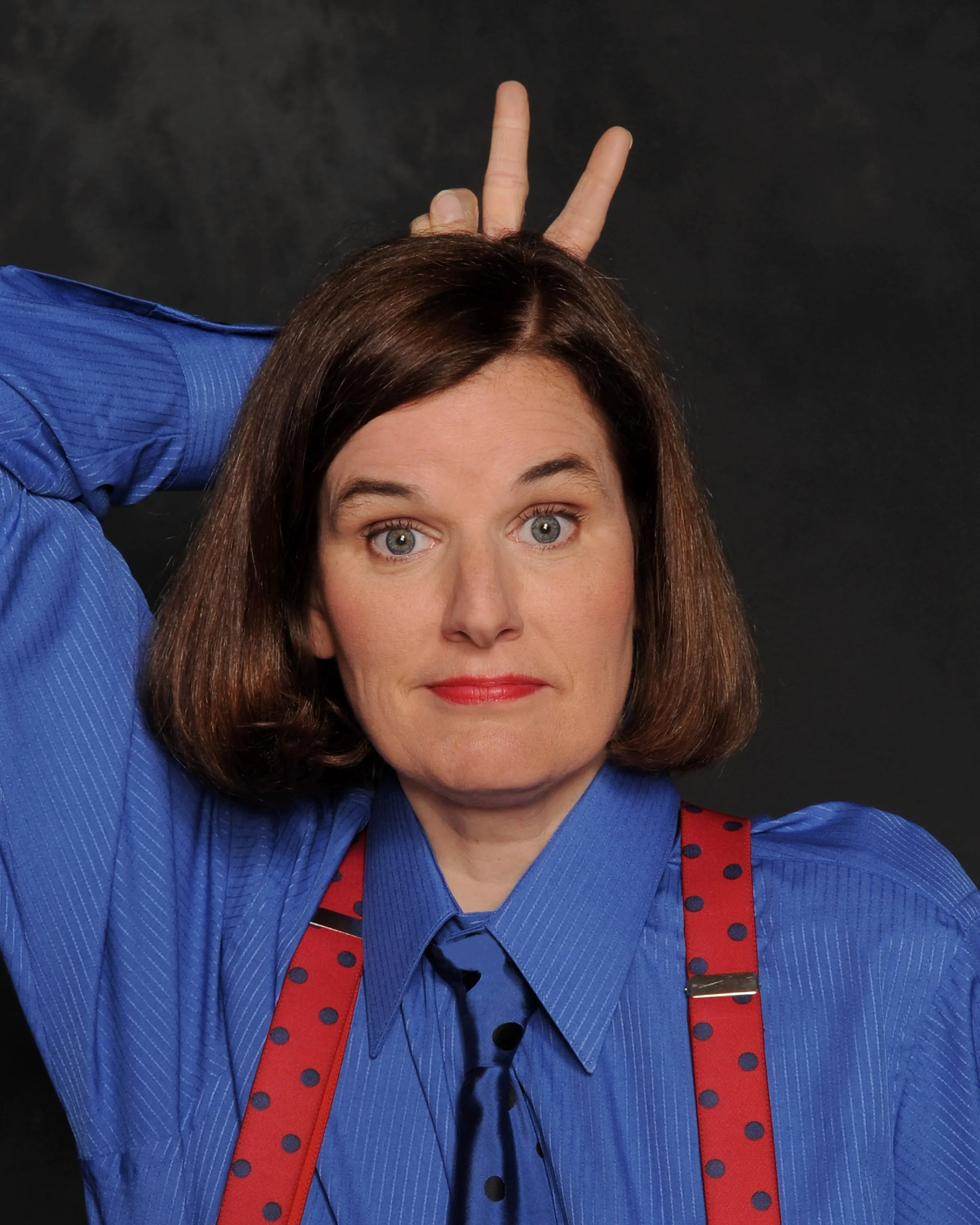 A woman with brown hair and blue eyes making a playful gesture with her hand behind her head, resembling bunny ears, wearing a blue shirt and red suspenders with polka dots, against a dark background.