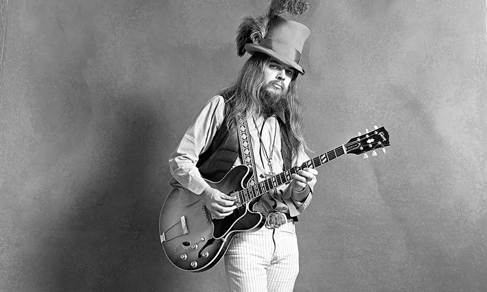 A man in a vintage hat with feathers, long hair, and a beard, holding a guitar, dressed in old-fashioned clothing, standing against a plain background.