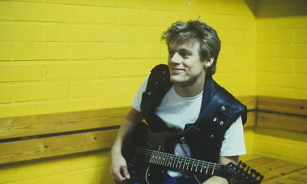 A young man with tousled hair, wearing a white t-shirt and a black vest, sitting on a wooden bench against a yellow brick wall, playing an electric guitar and smiling.