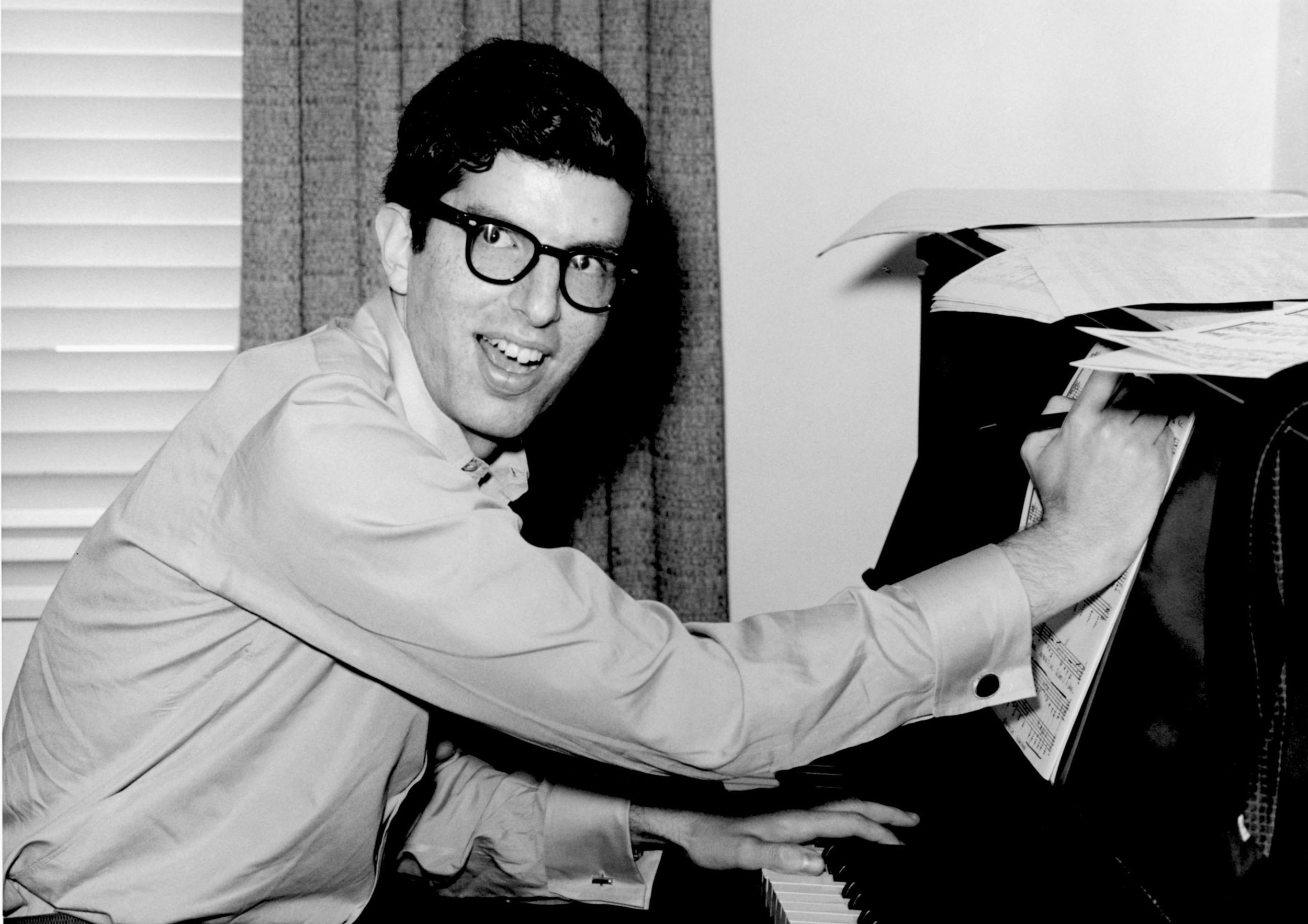 Black and white photo of a man with dark hair and glasses, smiling and playing the piano surrounded by scattered papers.