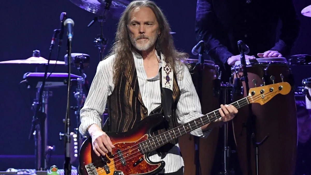 A man with long gray hair and a beard playing an electric bass guitar on stage, with percussion instruments in the background.