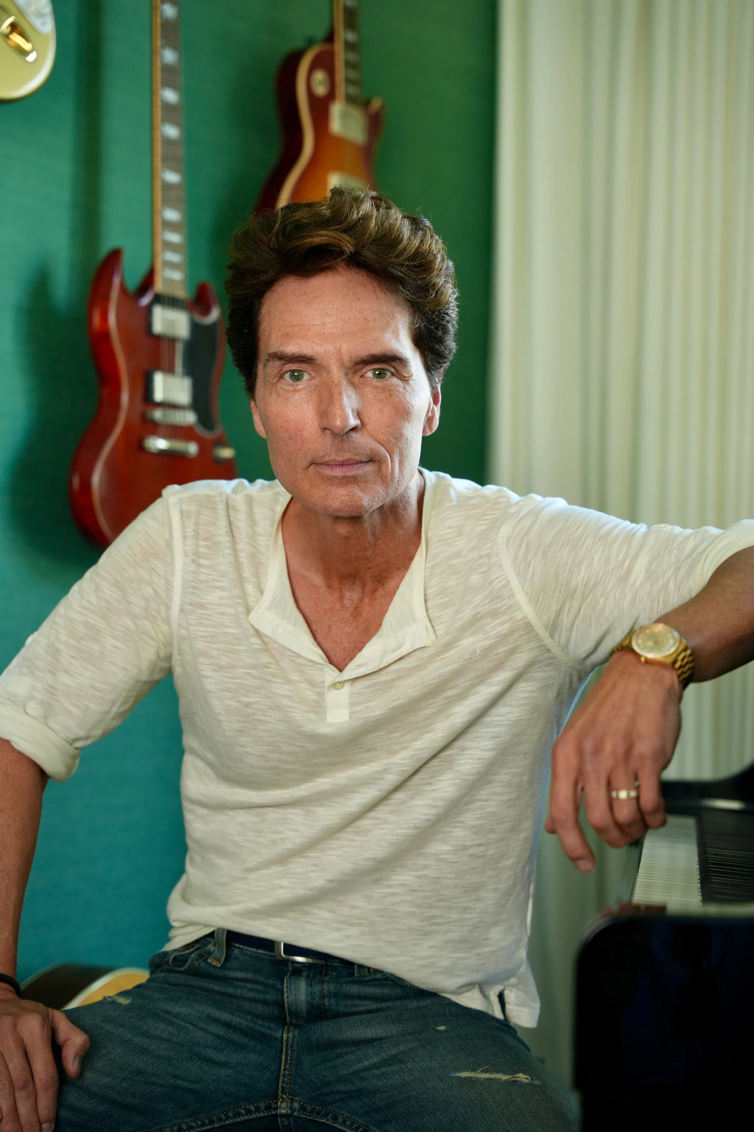 A man with dark, curly hair and light skin, wearing a white shirt and a gold wristwatch, sitting inside a room with green walls. Two electric guitars are hanging on the wall behind him, and a piano is partially visible on the right side of the image.