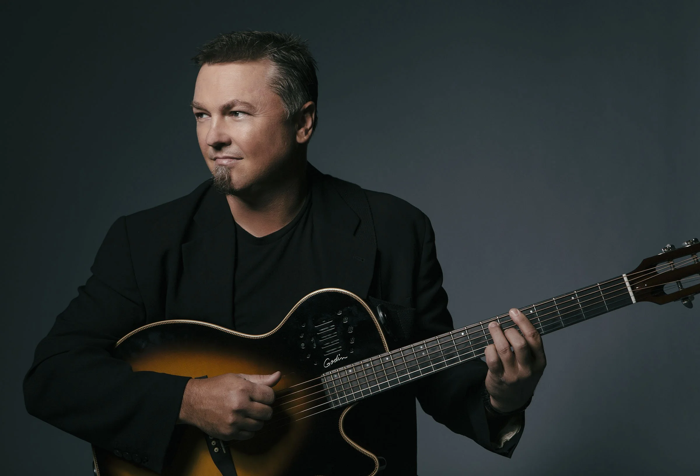 A man with short dark hair and a goatee, dressed in black, playing an acoustic guitar against a dark grey background.
