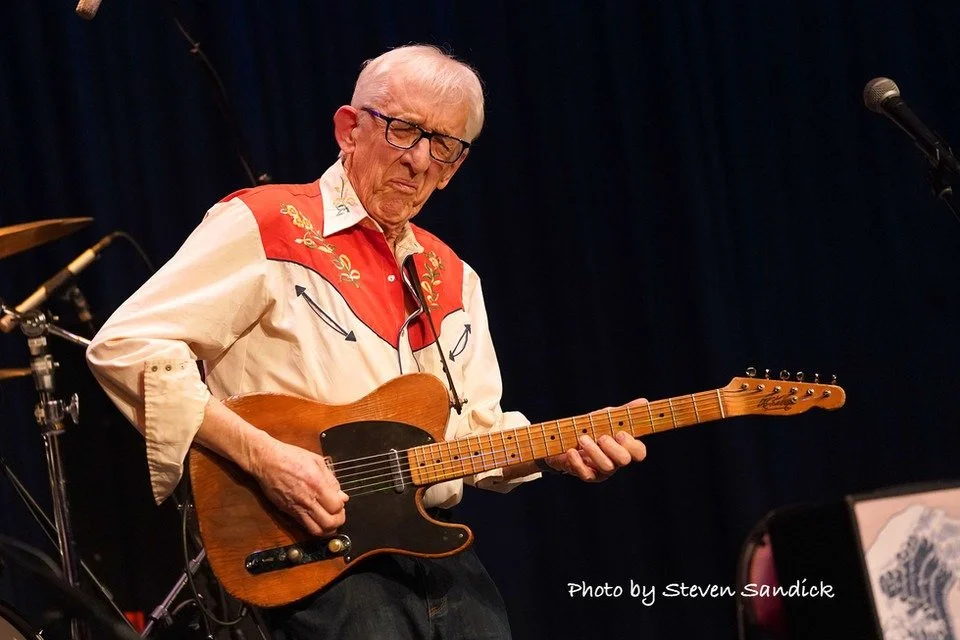 An elderly man playing a wooden electric guitar on stage. He wears glasses and a cowboy-style shirt with embroidery.