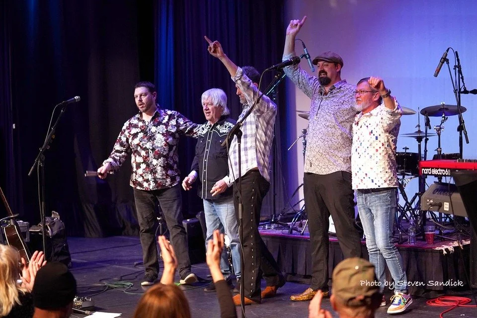 A group of five performers standing on a stage, taking a bow after a performance, with their arms around each other and fans clapping in the audience.