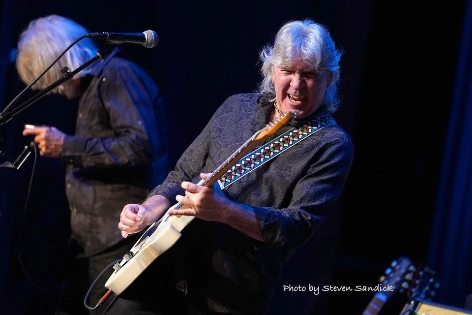 A male musician with long gray hair passionately playing an electric guitar on stage, with a woman in the background holding a microphone. The scene is lit with stage lights, and the background is dark.