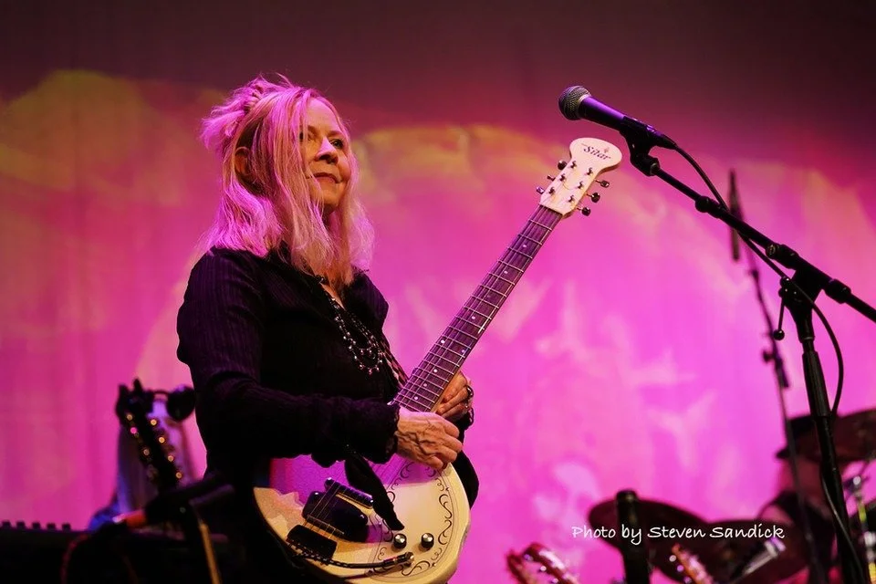 A woman with blonde hair holds a white electric guitar on stage, with a pink and purple background and a microphone setup nearby, during a performance.