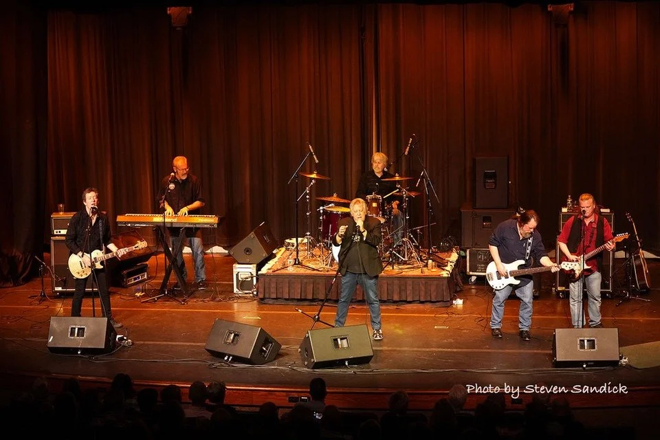 A six-member band performing on stage, with musicians playing guitars, drums, keyboard, and vocals, in front of a dark curtain.
