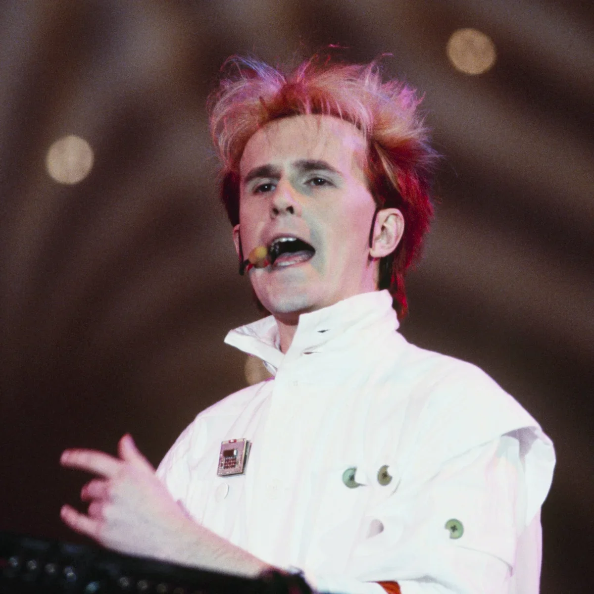 A performer with colorful hair and a white outfit singing or speaking on stage.