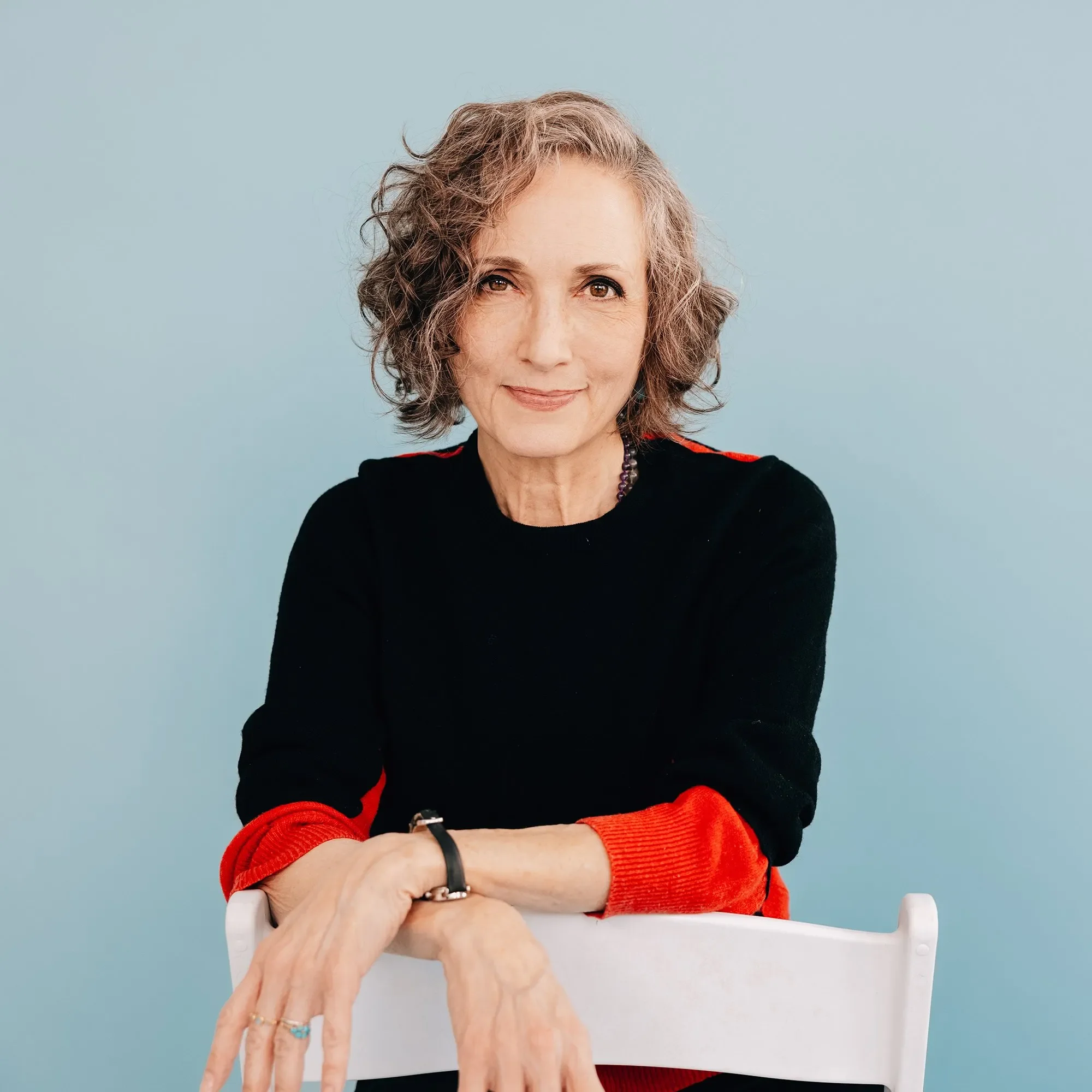 A woman with short, wavy gray hair smiling, wearing a black top with red sleeves, and sitting at a table with her arms crossed. An elderly person's hands are resting on her arm.