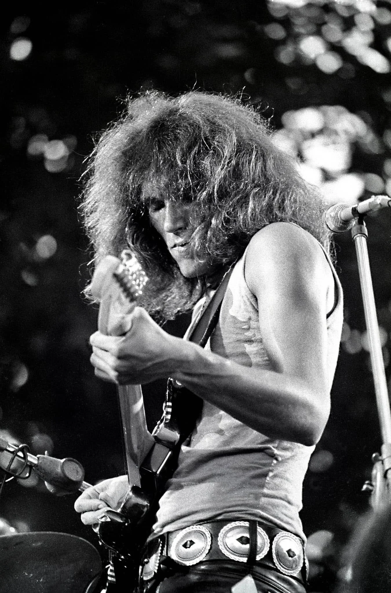 Black and white photo of a person with long hair playing an electric guitar on stage, with a music microphone, wearing a patterned skirt, and focused on the instrument.