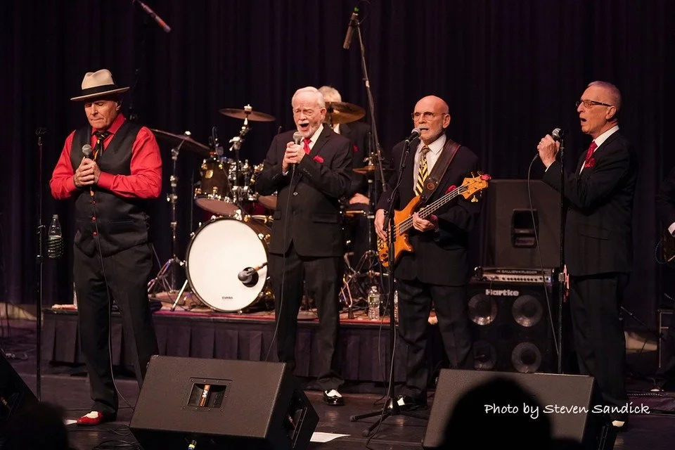 A band performing on stage with four male musicians singing into microphones, one playing bass, with drums and other equipment behind them.