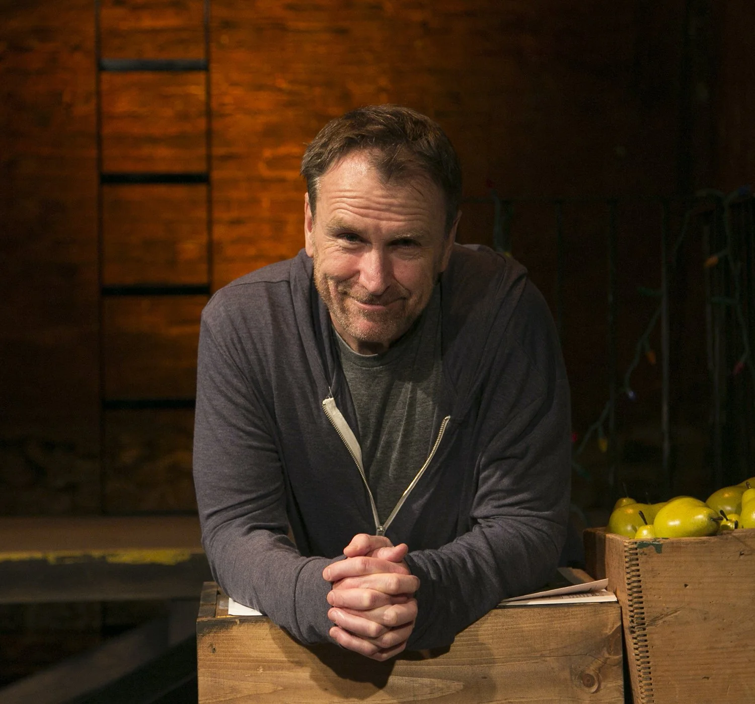 A middle-aged man with brown hair and a beard leaning on a wooden counter, smiling at the camera, behind a display of yellow tomatoes in a rustic setting.