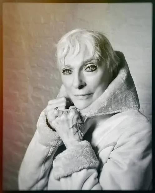 Black and white photo of an older woman with short hair, wearing a light-colored jacket with a fur-lined collar, posing against a plain background.