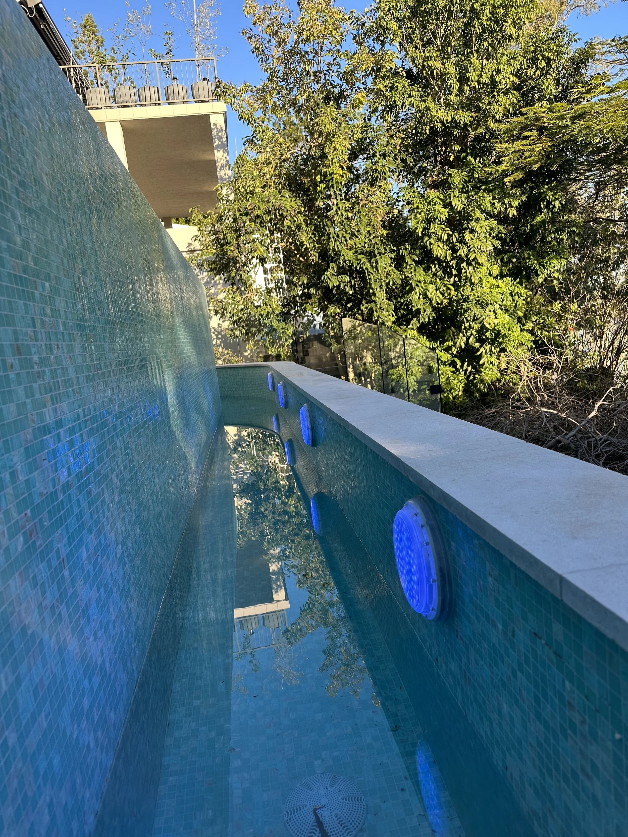 View of a swimming pool with a water slide and blue LED lights on the side, overlooking trees and a building with an outdoor balcony and chairs.