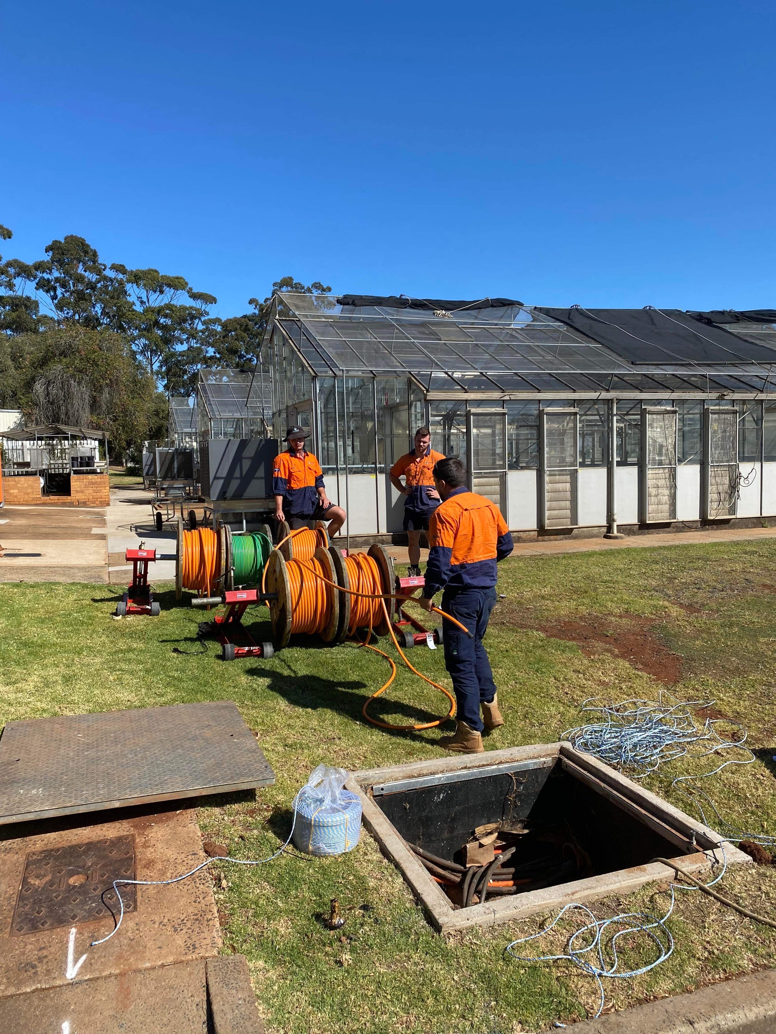 Workers in orange and black uniforms working outside near a greenhouse, with various equipment, hoses, and open manholes on the ground under a clear blue sky.
