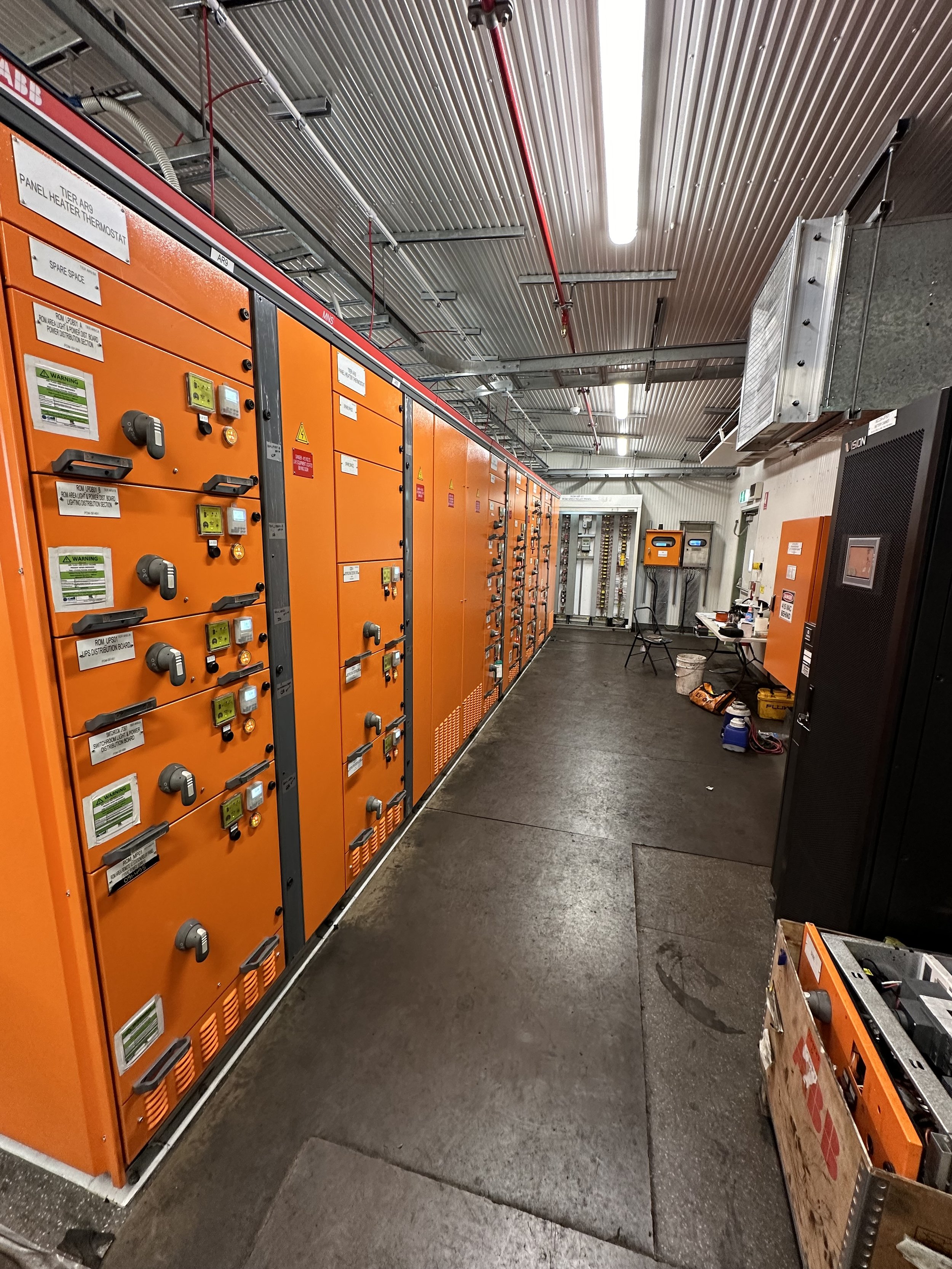 An industrial electrical room with orange electrical panels lined up along the wall, various warning labels, and control switches. The ceiling has metal panels with red pipes and fluorescent lighting. The floor is dark and slightly wet, with tools an