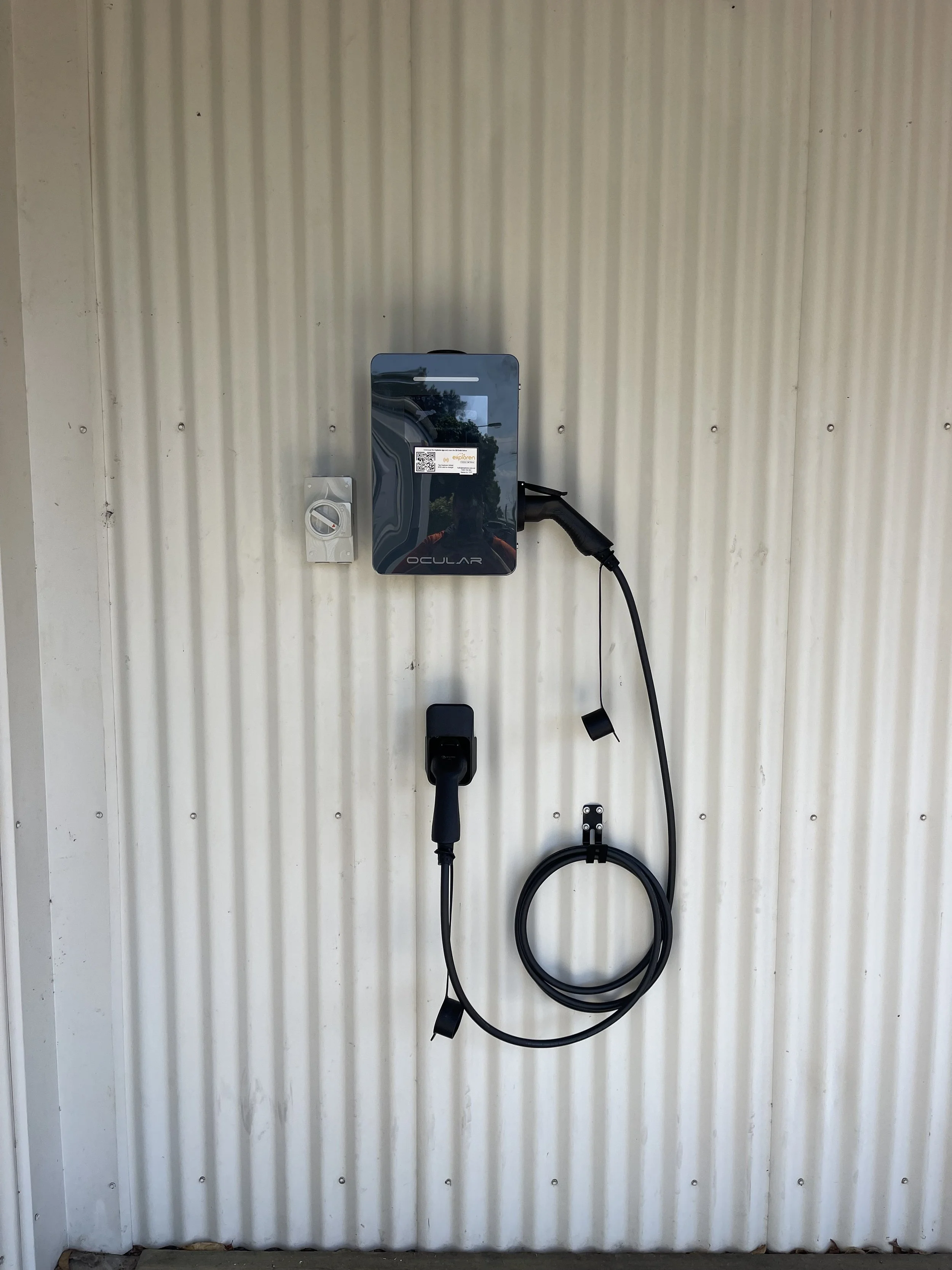 Electric vehicle charging station with a charger plugged into an electric car wall port, mounted on a corrugated metal wall.