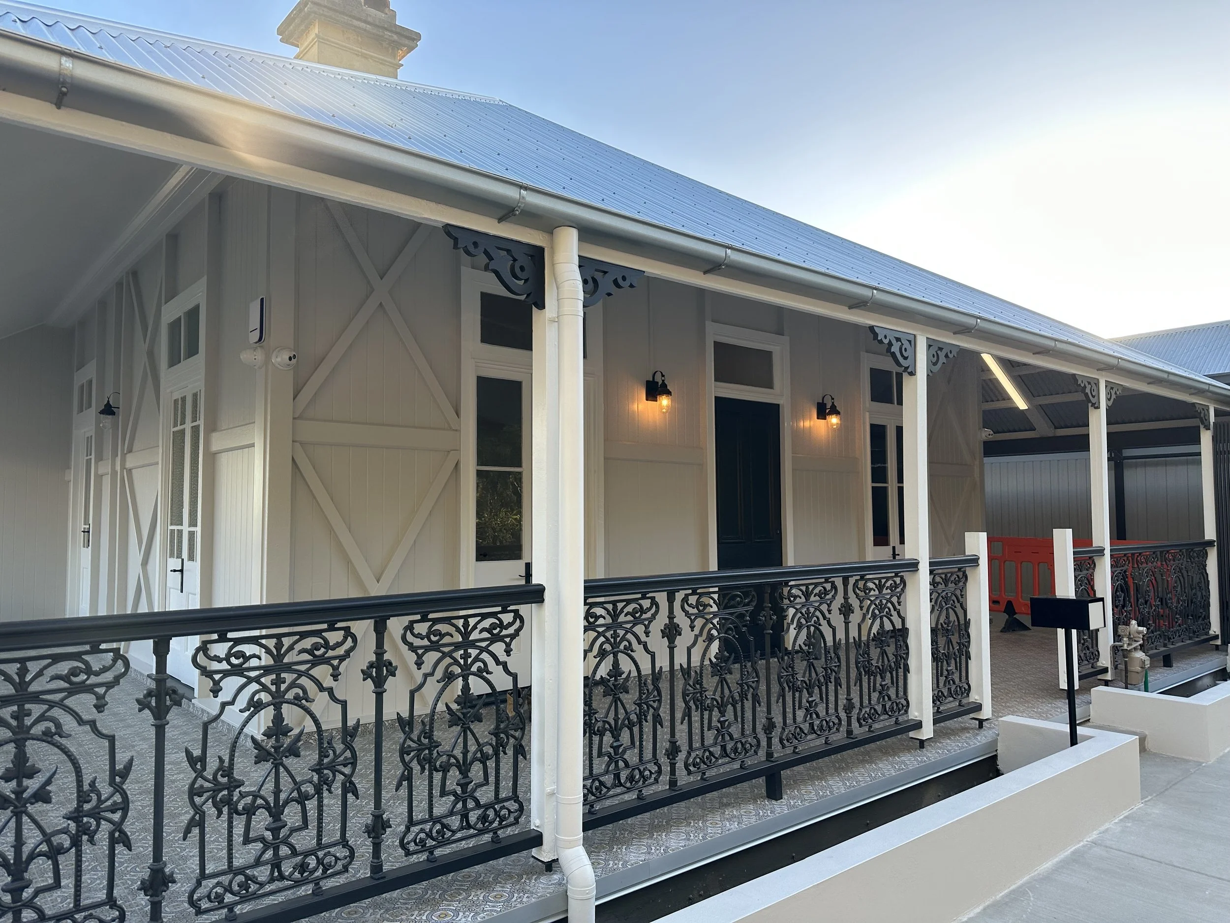 Exterior view of a building with decorative black wrought iron balcony railing, white siding, large windows, and black outdoor lighting fixtures, with a metal roof and clear sky in the background.