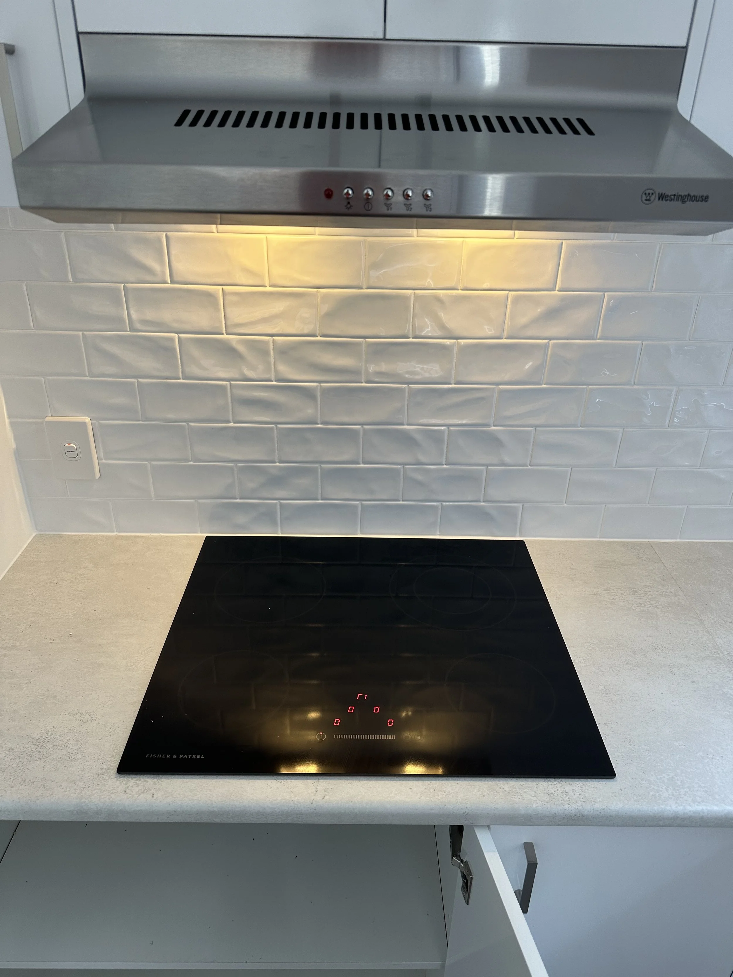 Stainless steel kitchen range hood above a black electric cooktop on a white countertop, with tiled backsplash.