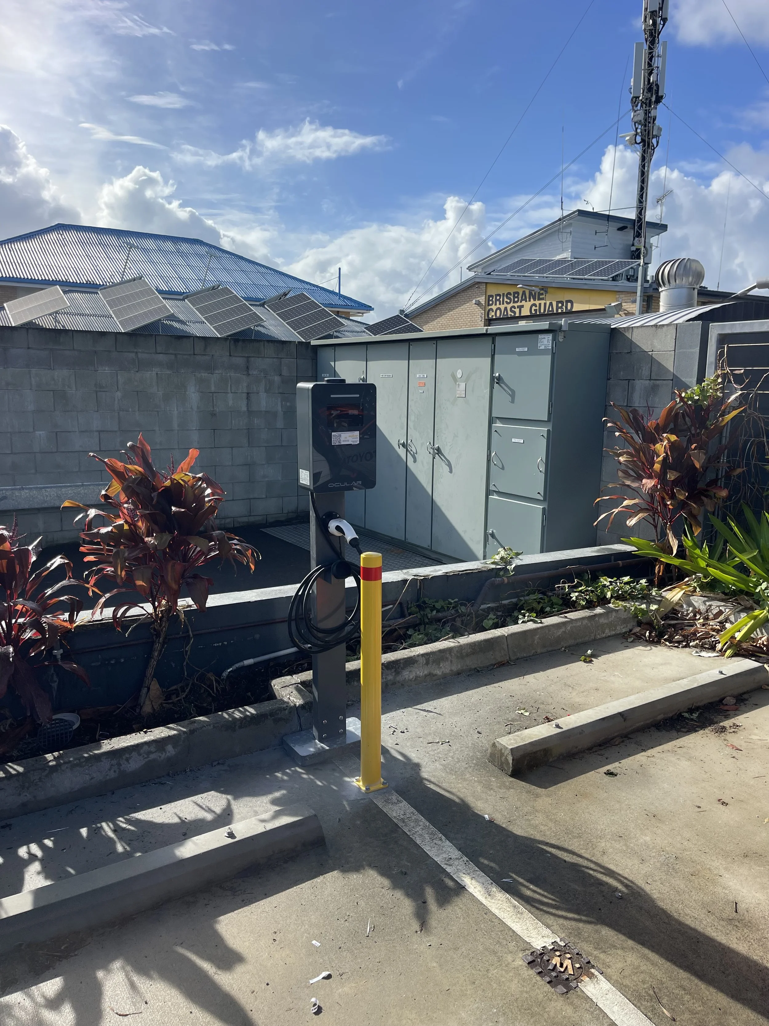 Electric vehicle charging station in parking lot, with empty parking spaces and a concrete block wall, building with solar panels and a sign reading 'Brisbane Coast Guard' in the background.