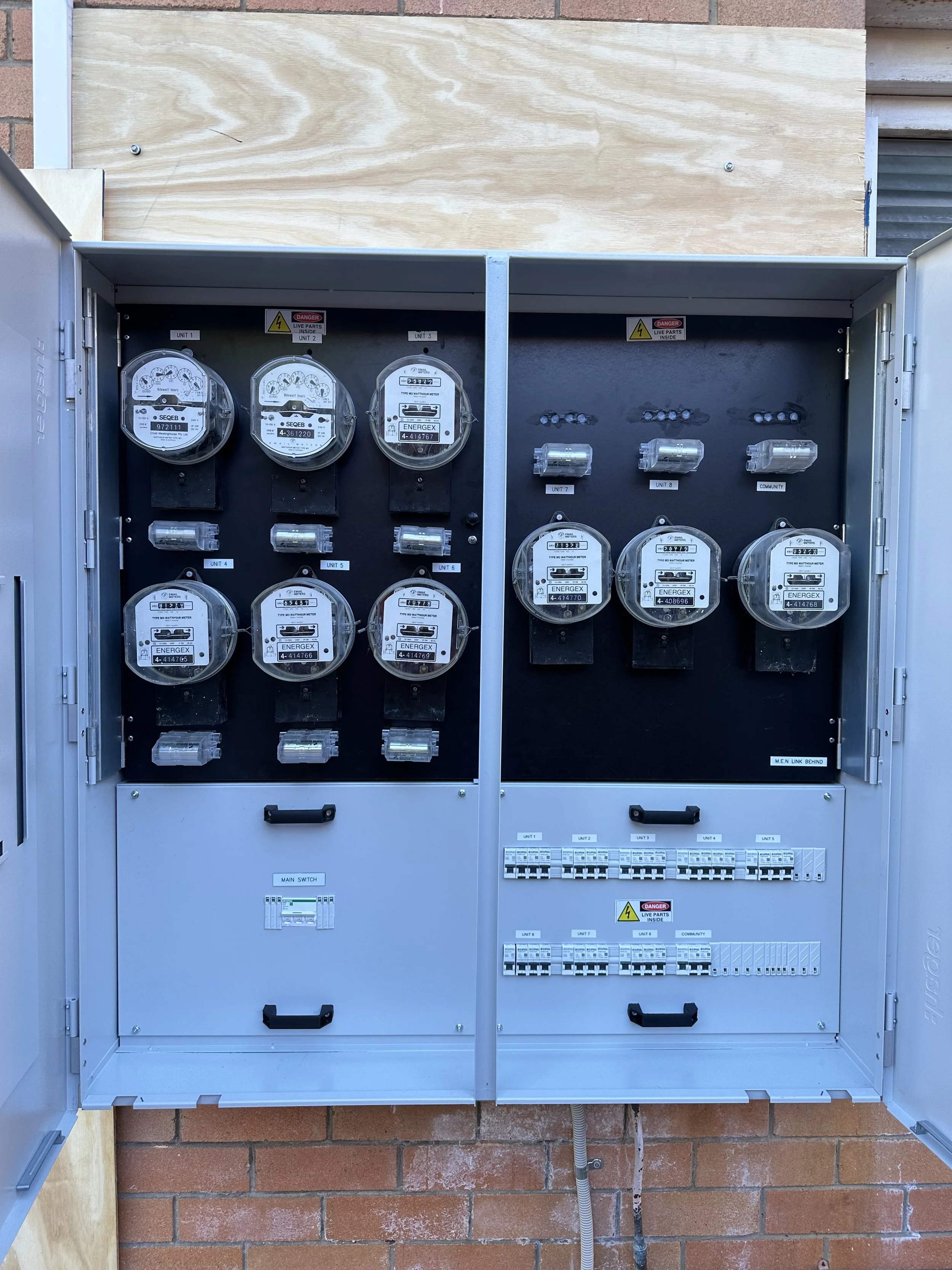 An electrical utility box with multiple meters, circuit breakers, and warning labels, mounted on a brick wall behind a wooden board.