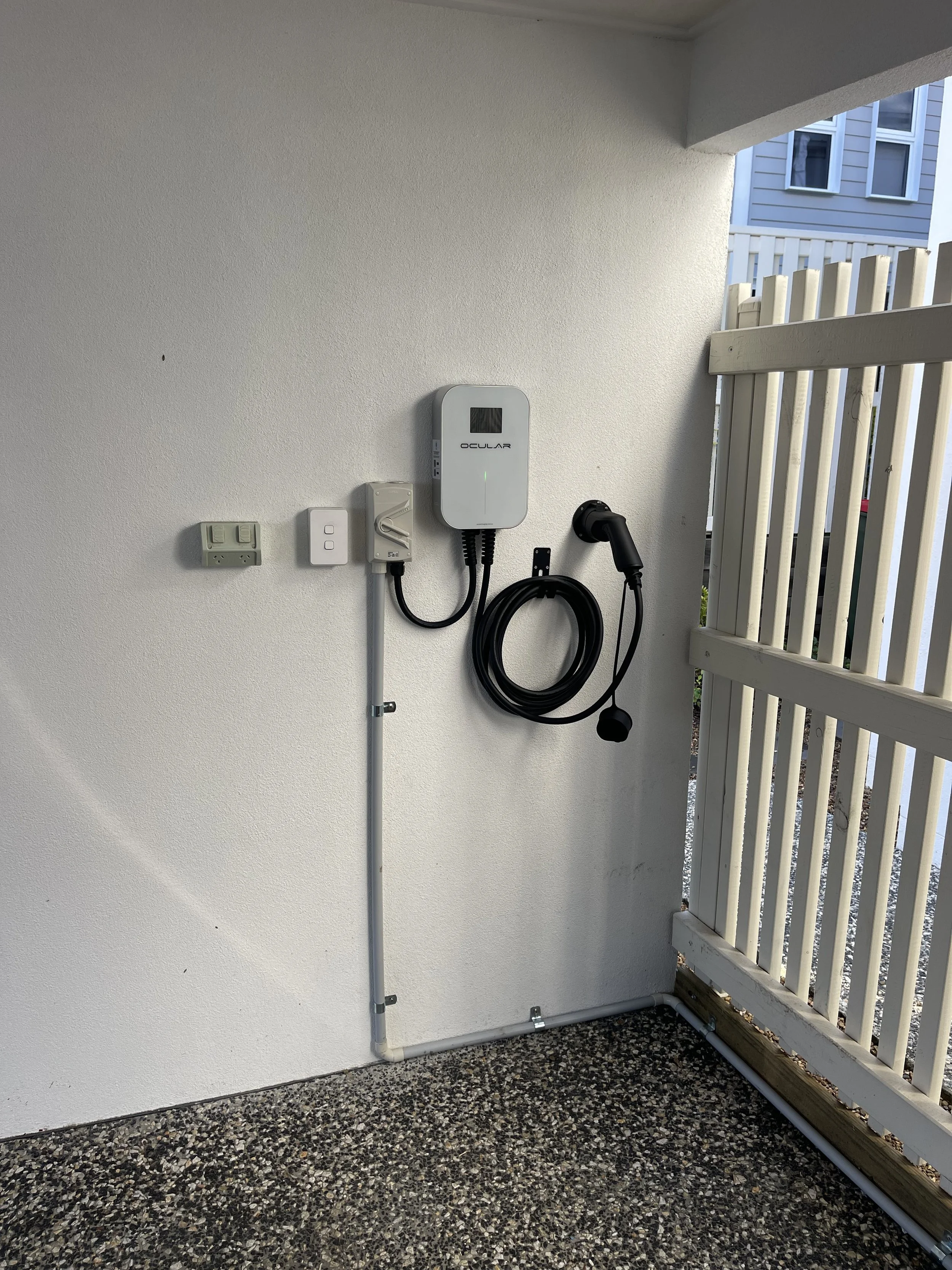 A small outdoor area with a white wall, electrical outlets, a charging station, and an electric vehicle charging cable. There is a white wooden fence on the right side, and it appears to be part of a residential nearby building.