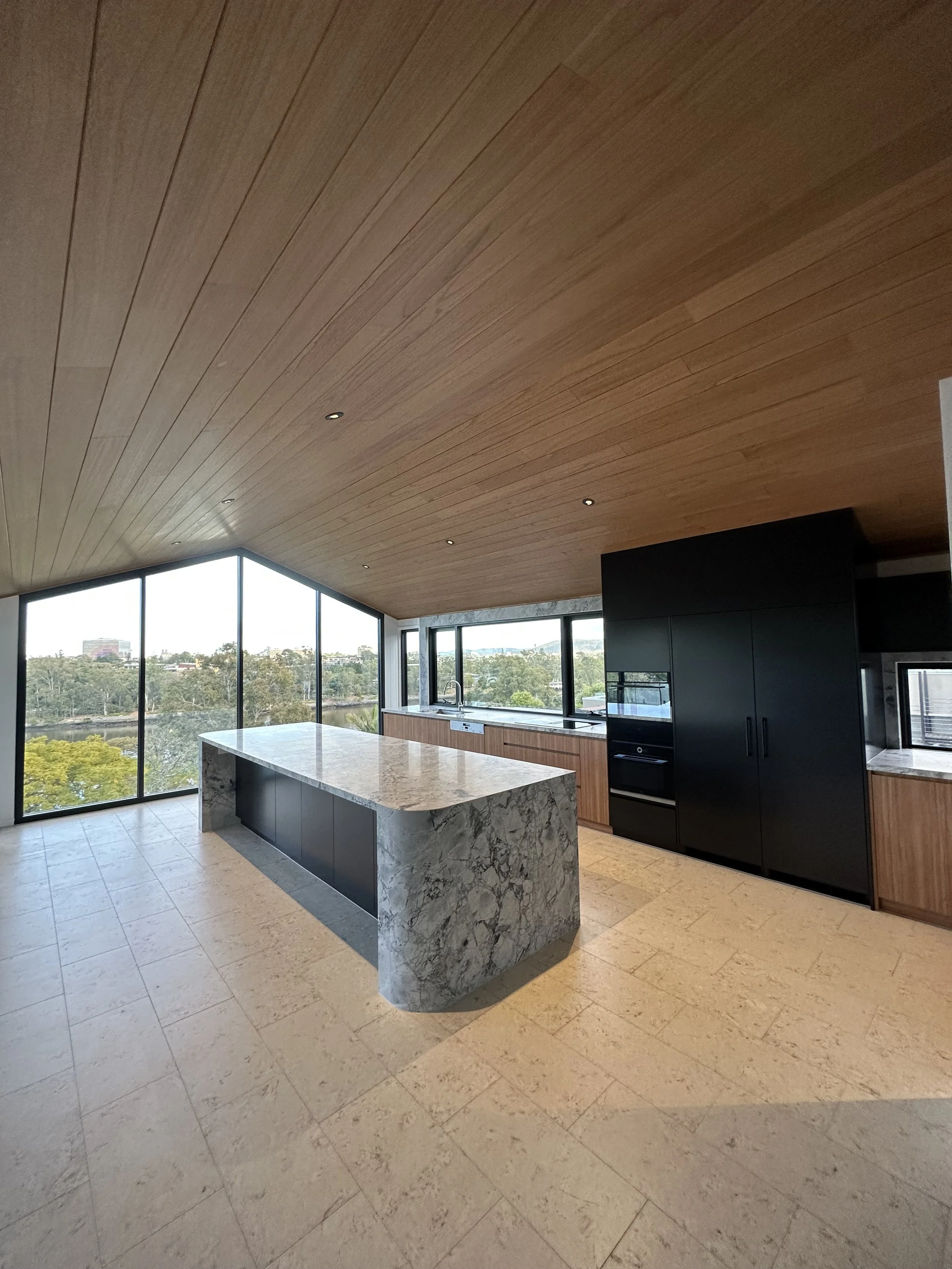 Modern kitchen with large marble island, black cabinetry, wooden ceiling, and floor-to-ceiling windows showing an outdoor view.