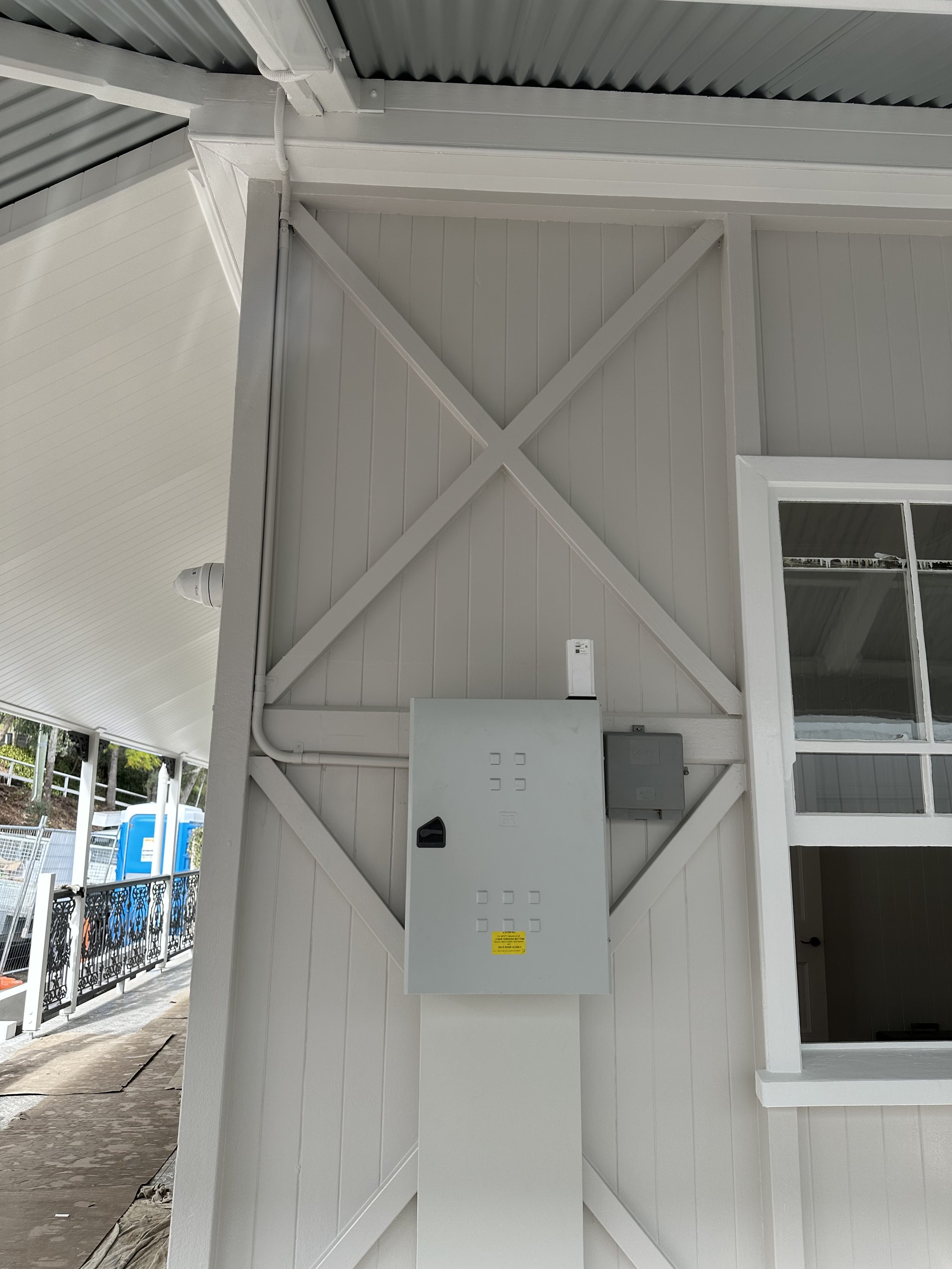 Close-up of a corner of a white corrugated metal roof with an electrical box mounted on the wall underneath, electrical wires and conduit running along the corner, and a window with a partial view of the outdoor area.