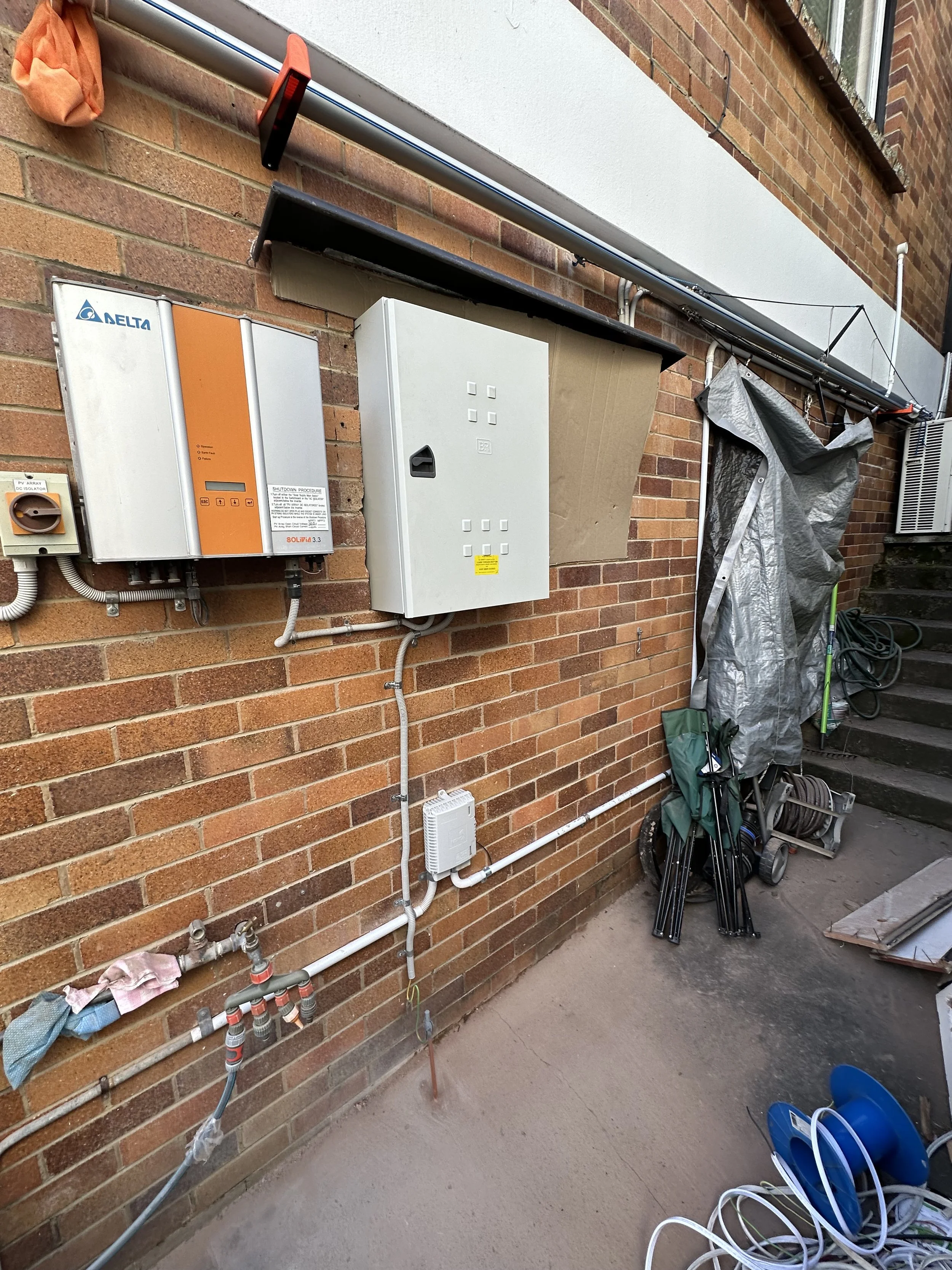 Outdoor wall with electrical boxes, pipes, and gardening tools, near stairs and a window.