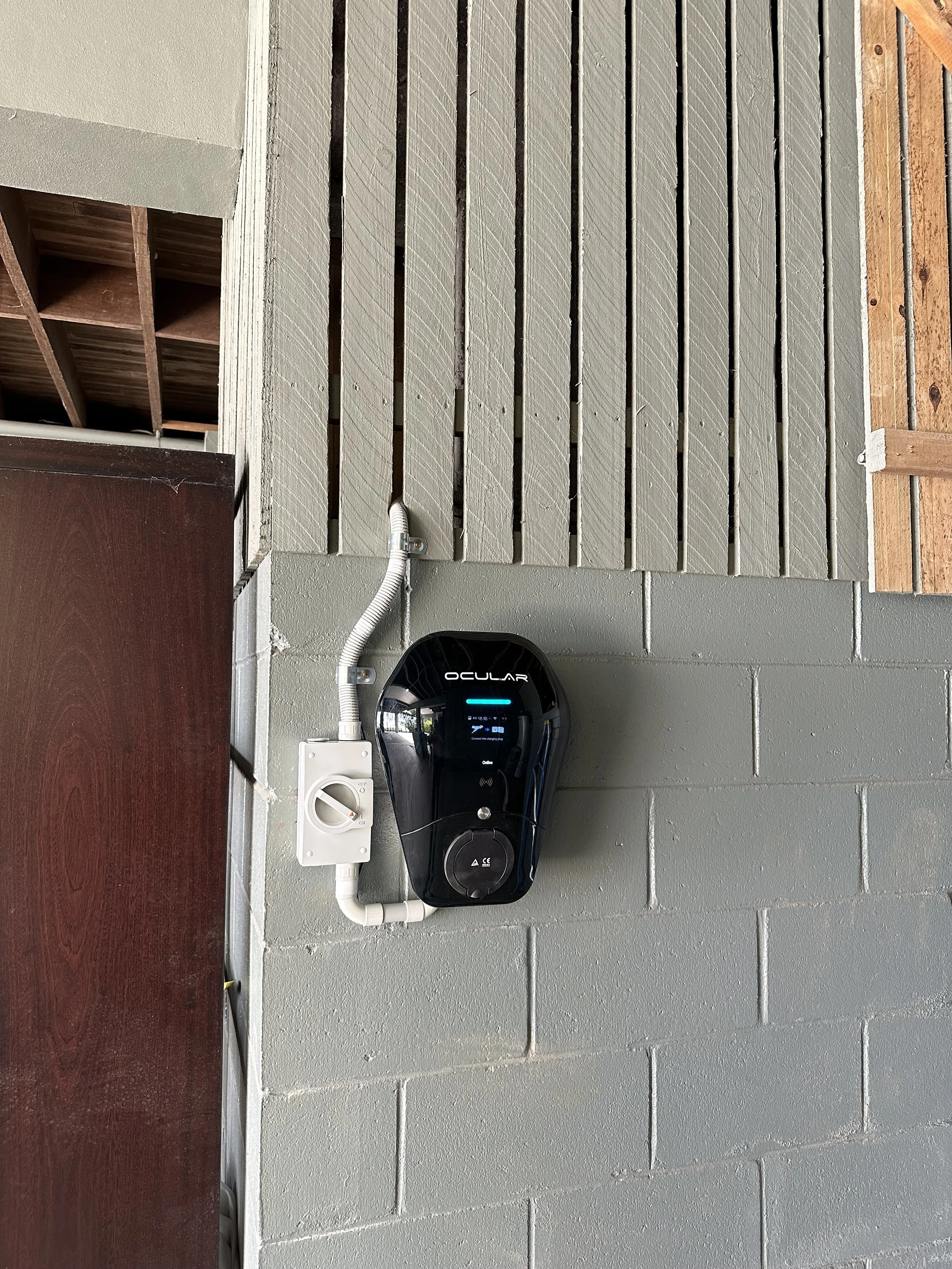 Close-up of an electric vehicle charging station mounted on a gray brick wall, with white electrical conduit and a power outlet nearby.