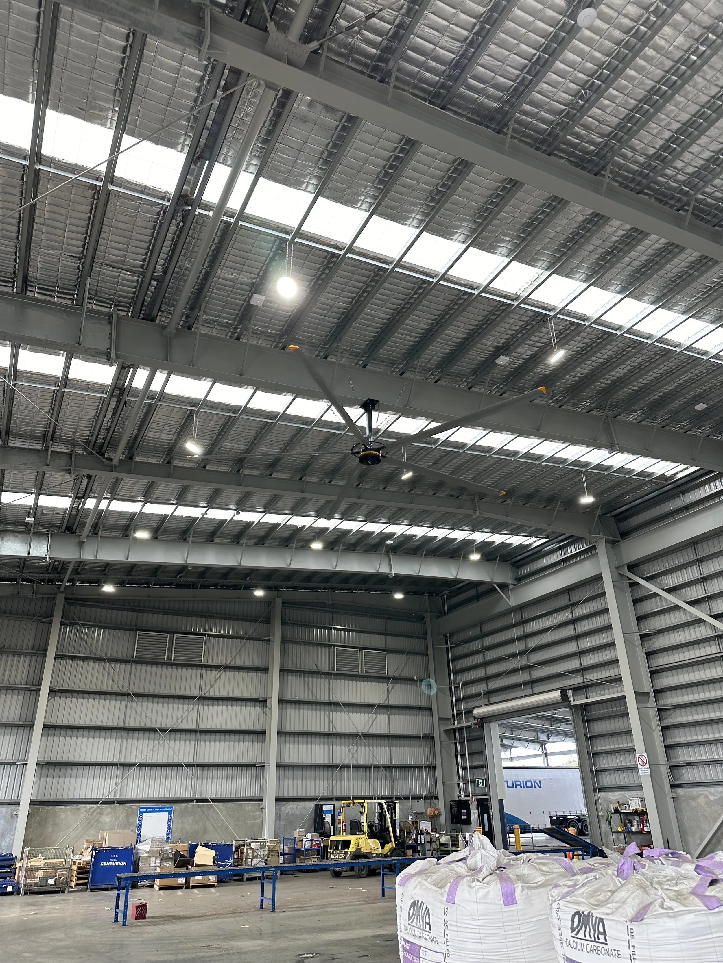 Interior view of a large industrial warehouse under construction, with construction materials, boxes, a forklift, and large bags of calcium carbonate on the floor.