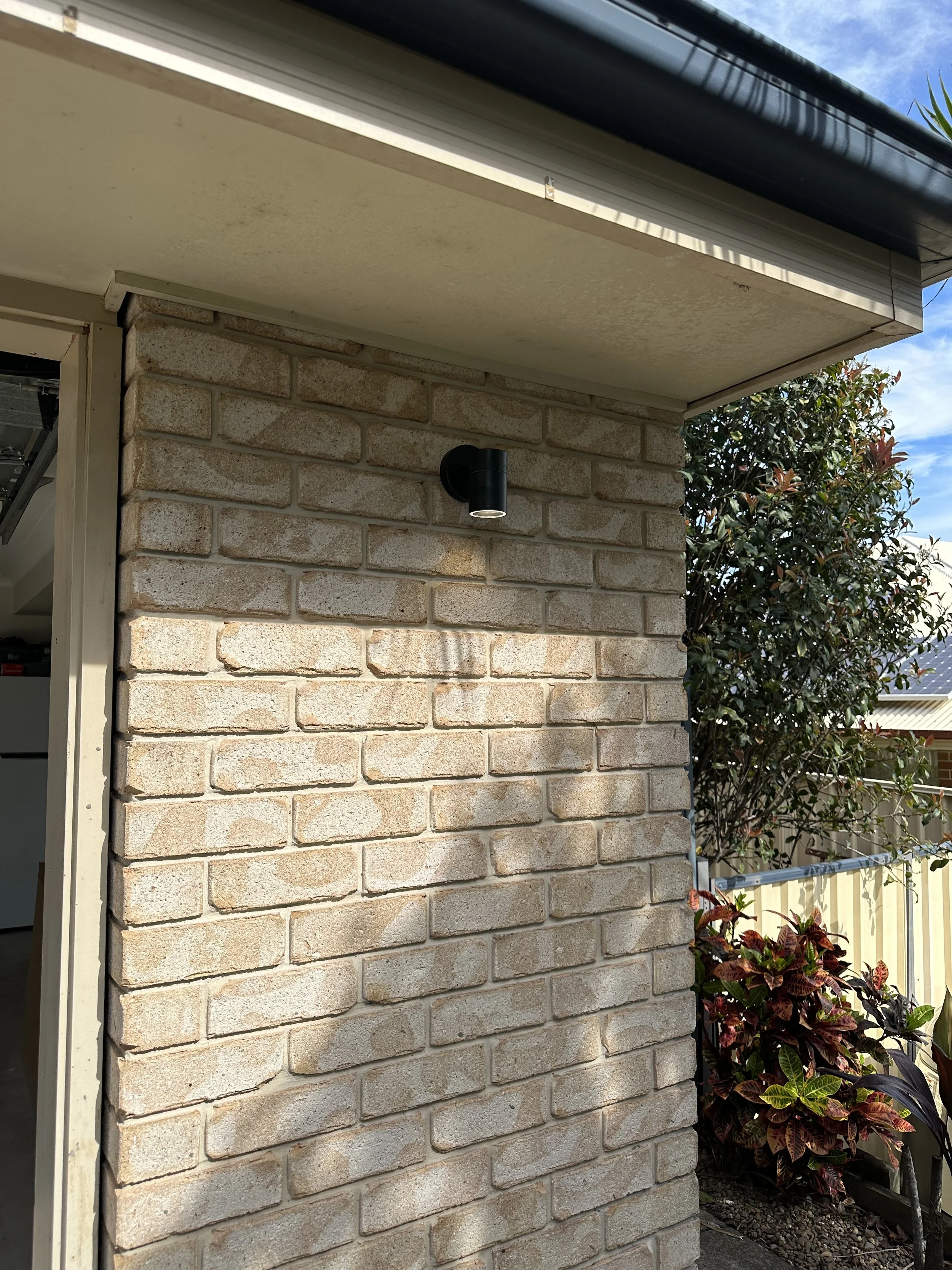 Exterior corner of a brick house with an outdoor light fixture and a small garden with plants along the side.