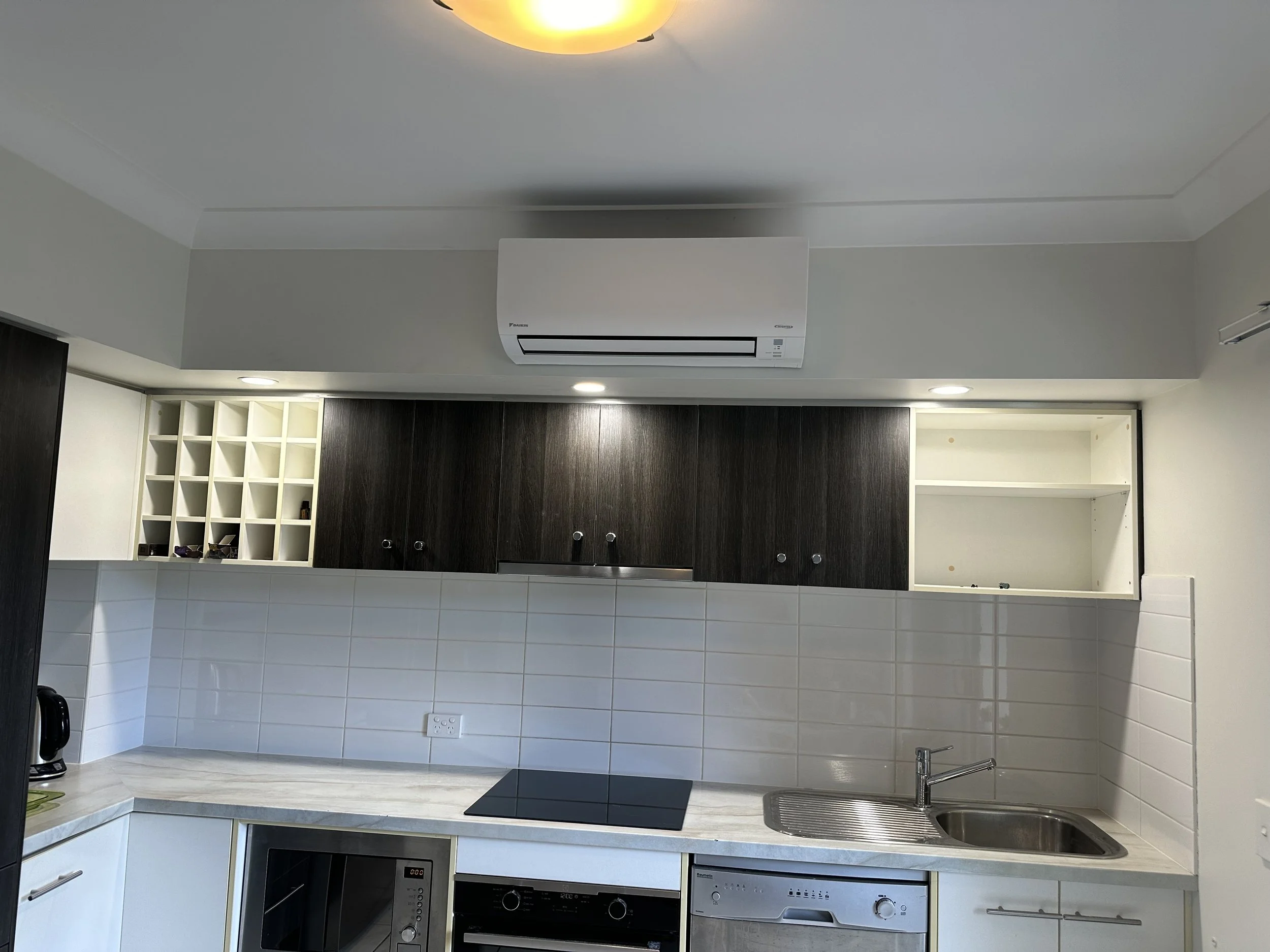 Modern kitchen with white tiled backsplash, white and dark wood cabinetry, a built-in oven, a stovetop, a sink, and upper cabinets. There is a wine rack in the upper cabinets and various minimal kitchen items on the counter.