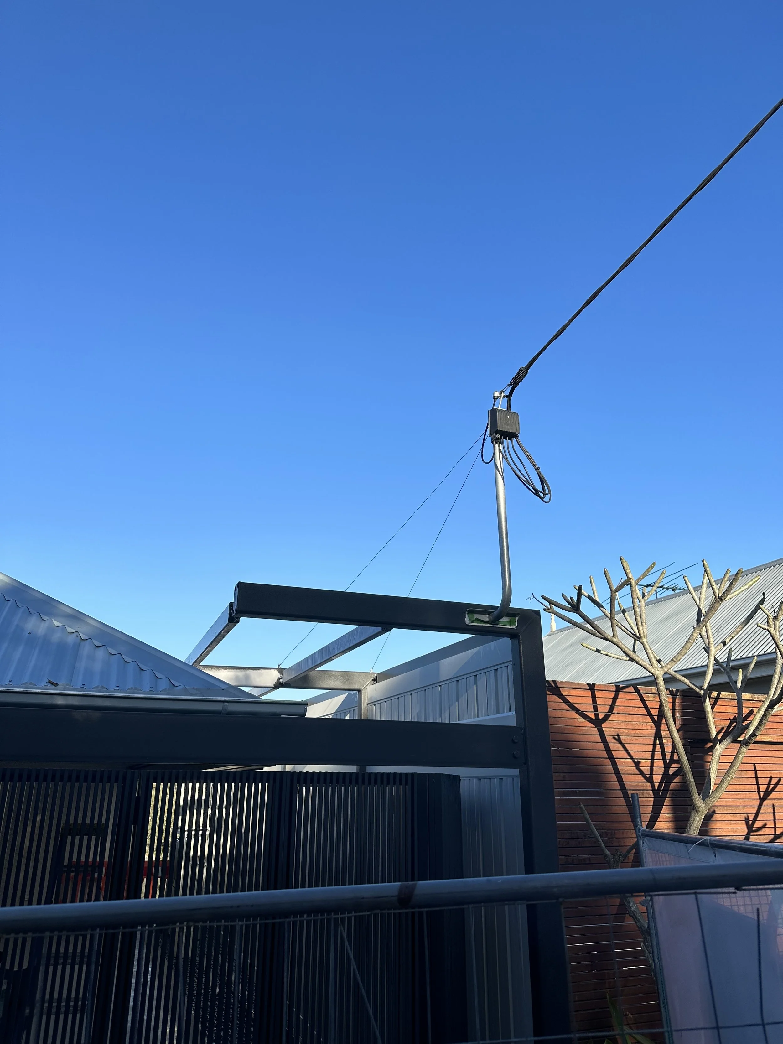A tall metal pole with electrical wires and a transformer, set against a clear blue sky. There are rooftops and a leafless tree in the background.