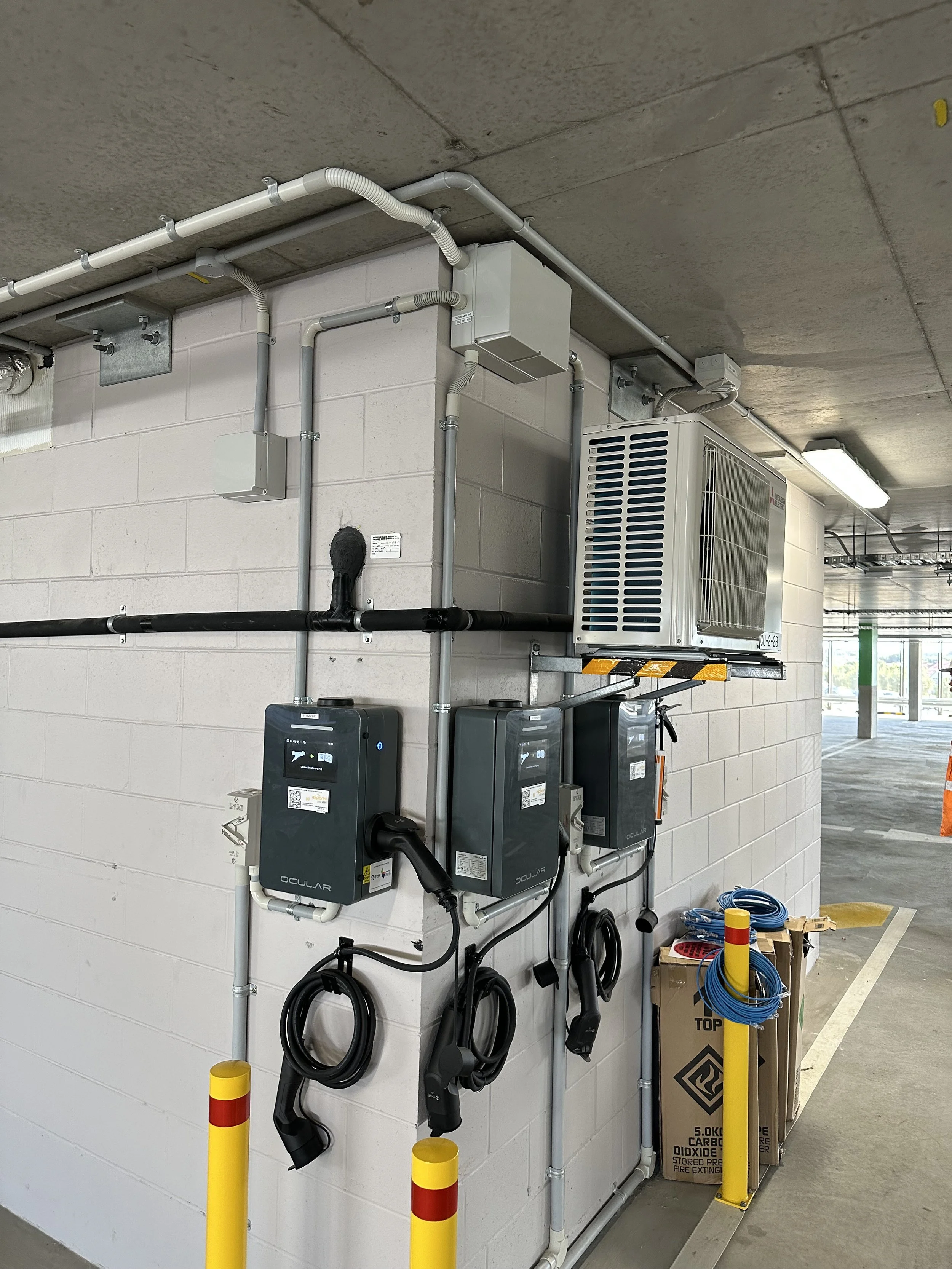 Electrical and plumbing equipment mounted on a white brick wall in a parking garage or industrial space, with yellow safety bollards in the foreground.
