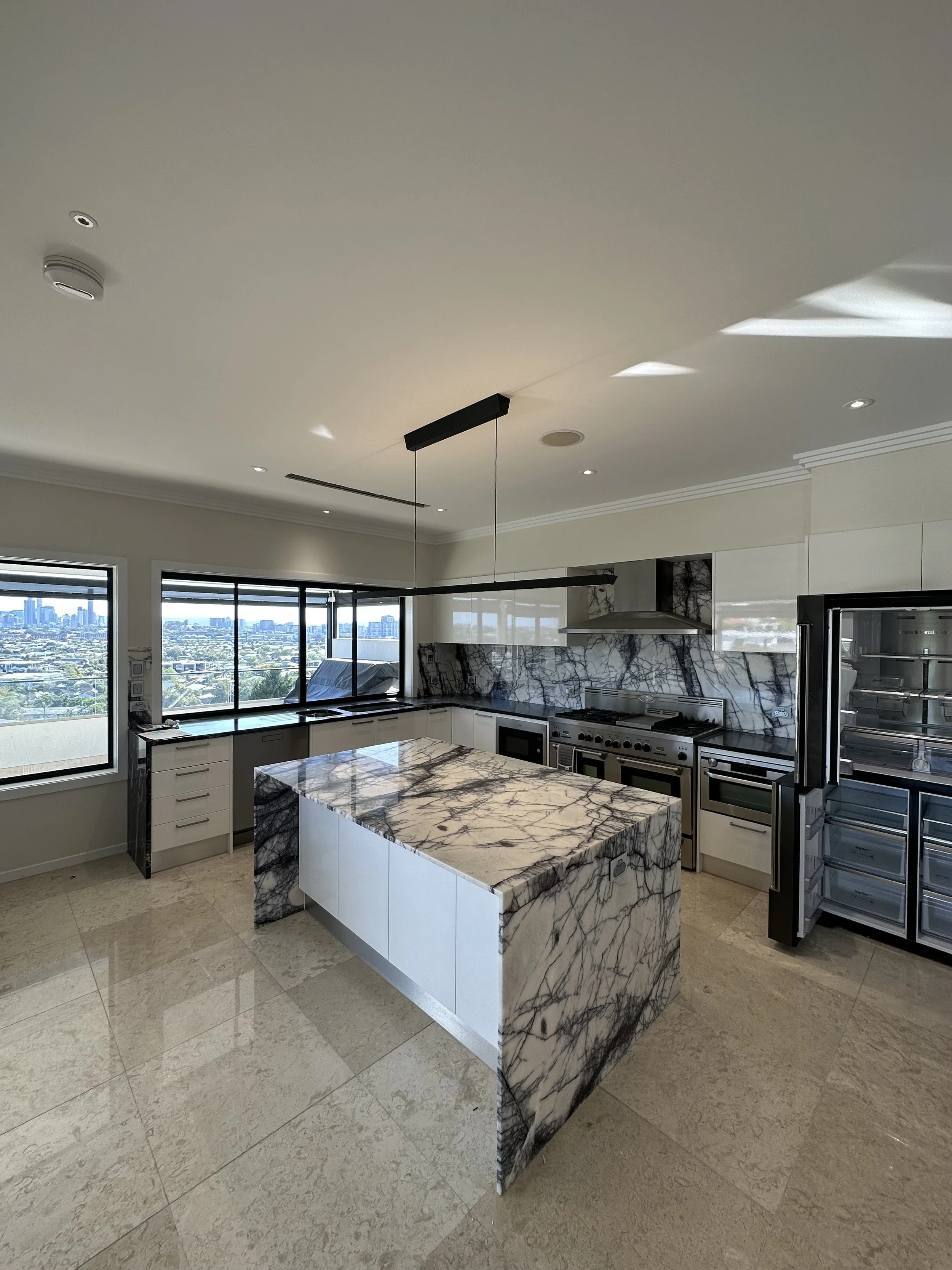 Modern kitchen with marble countertops and island, stainless steel appliances, large windows, city skyline in the distance, neutral color scheme.