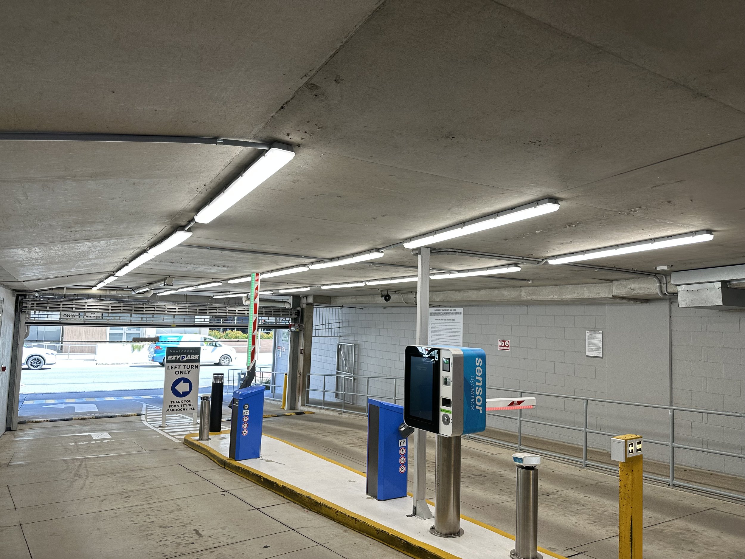 Parking garage entry with barrier gate, payment terminal, and signs indicating left turn only for exit, with cars and a building visible outside.