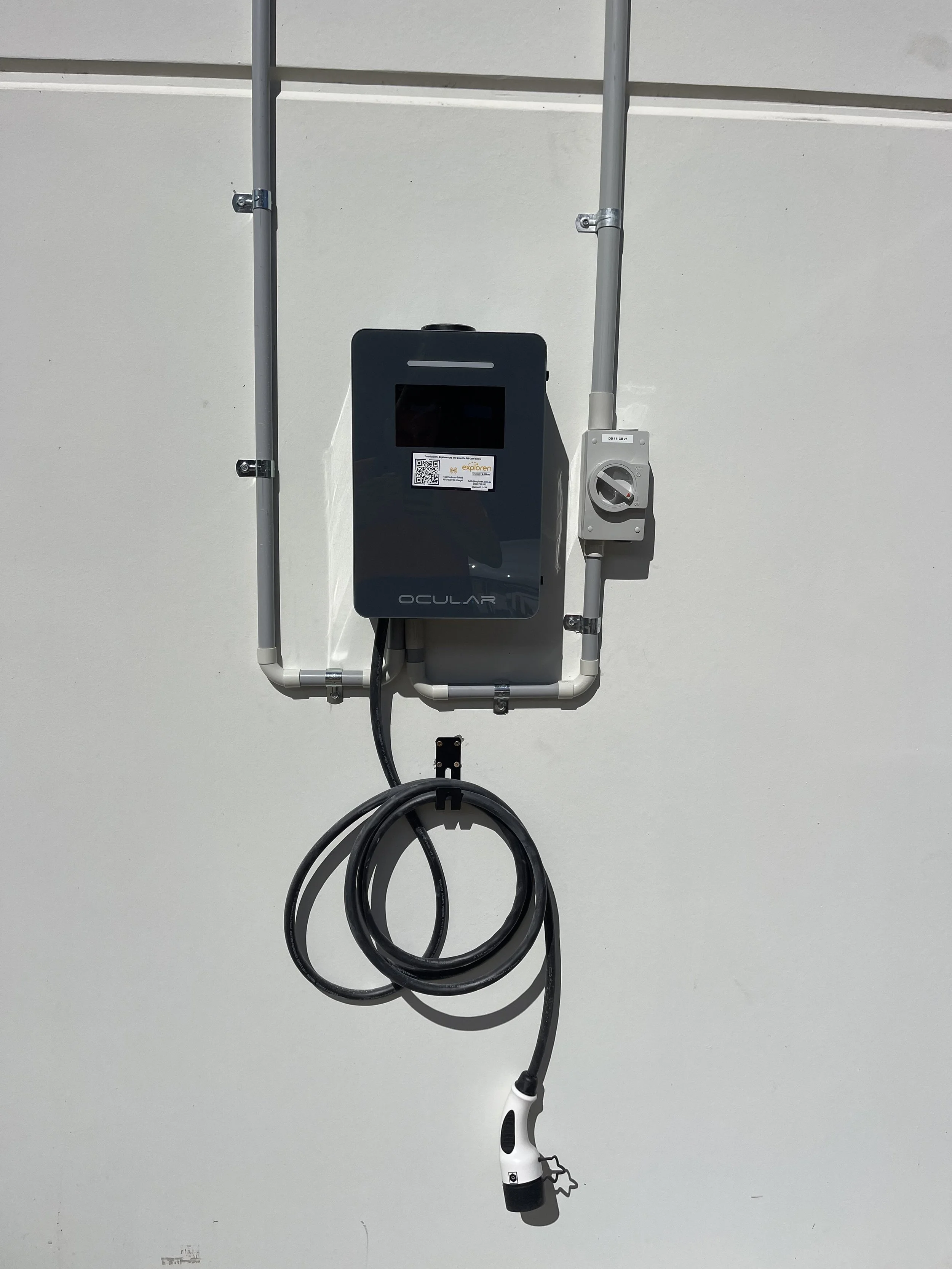 Electric vehicle charging station installed on a wall with exposed pipes, featuring a black screen, a QR code sticker, and a white charging plug hanging from a coiled cable.